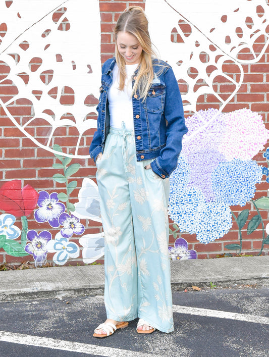 Woman in a denim jacket and light blue pants standing in front of a colorful mural on a brick wall.