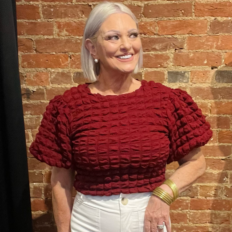 A woman standing against a brick wall, wearing a red textured stretchy top with puff sleeves and a classic crew neckline, paired with white pants.
