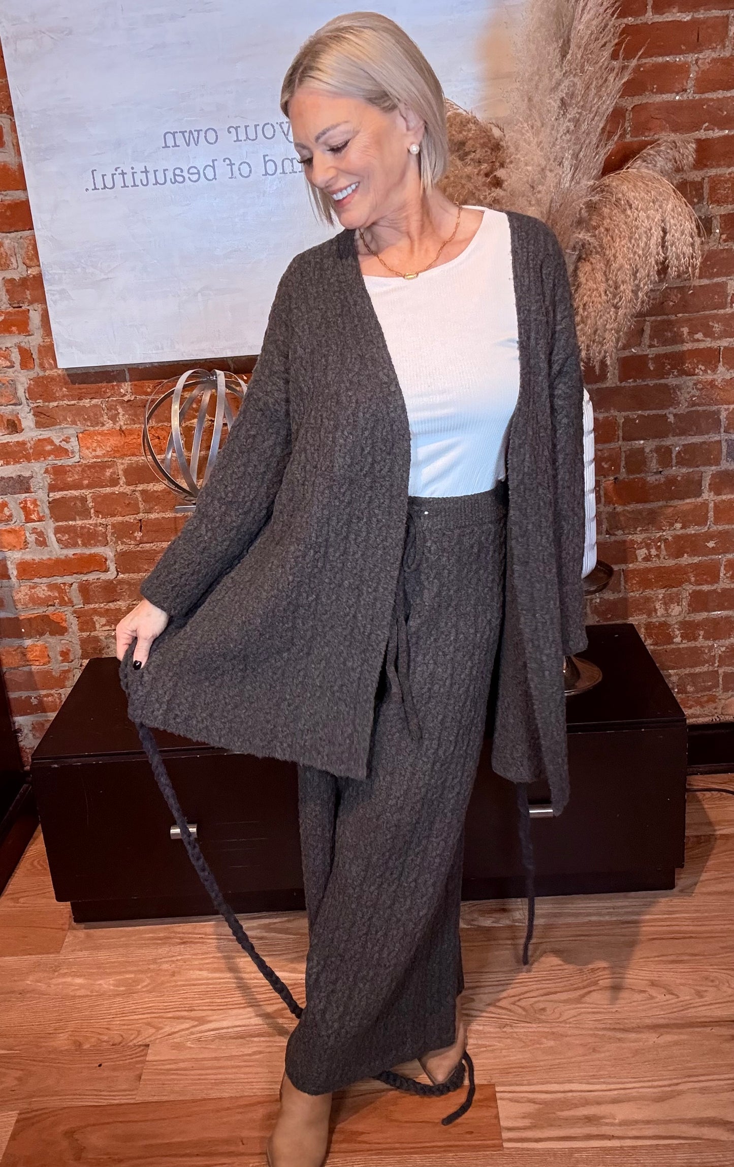Woman wearing a gray cardigan and pants standing in front of a brick wall.