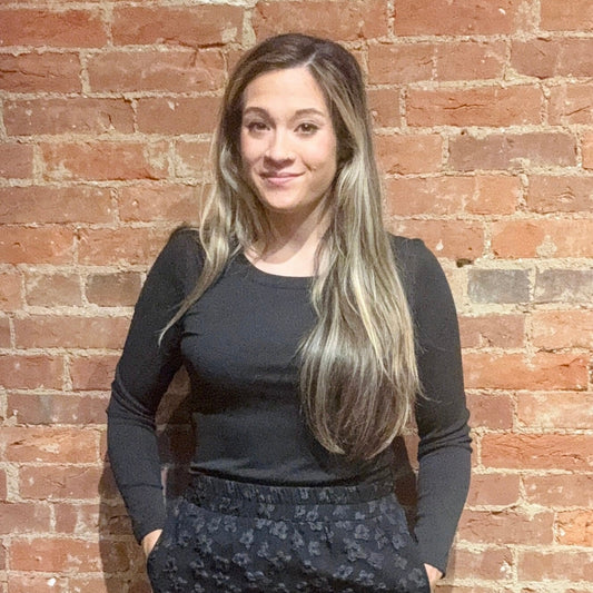 Woman wearing a black long-sleeve top and patterned pants standing against a brick wall.