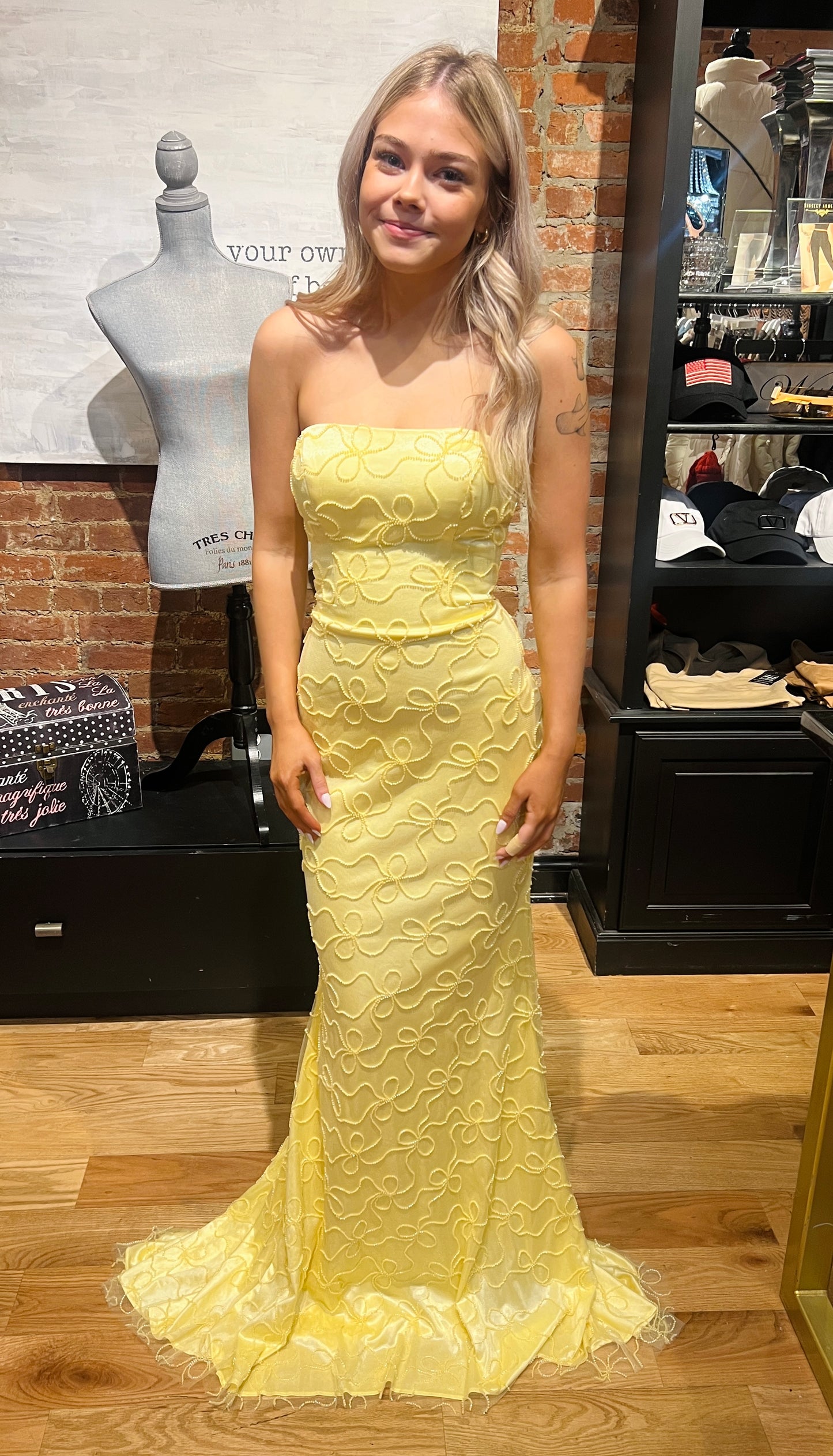Woman wearing a yellow strapless dress in an indoor setting with mannequins and clothing displays.