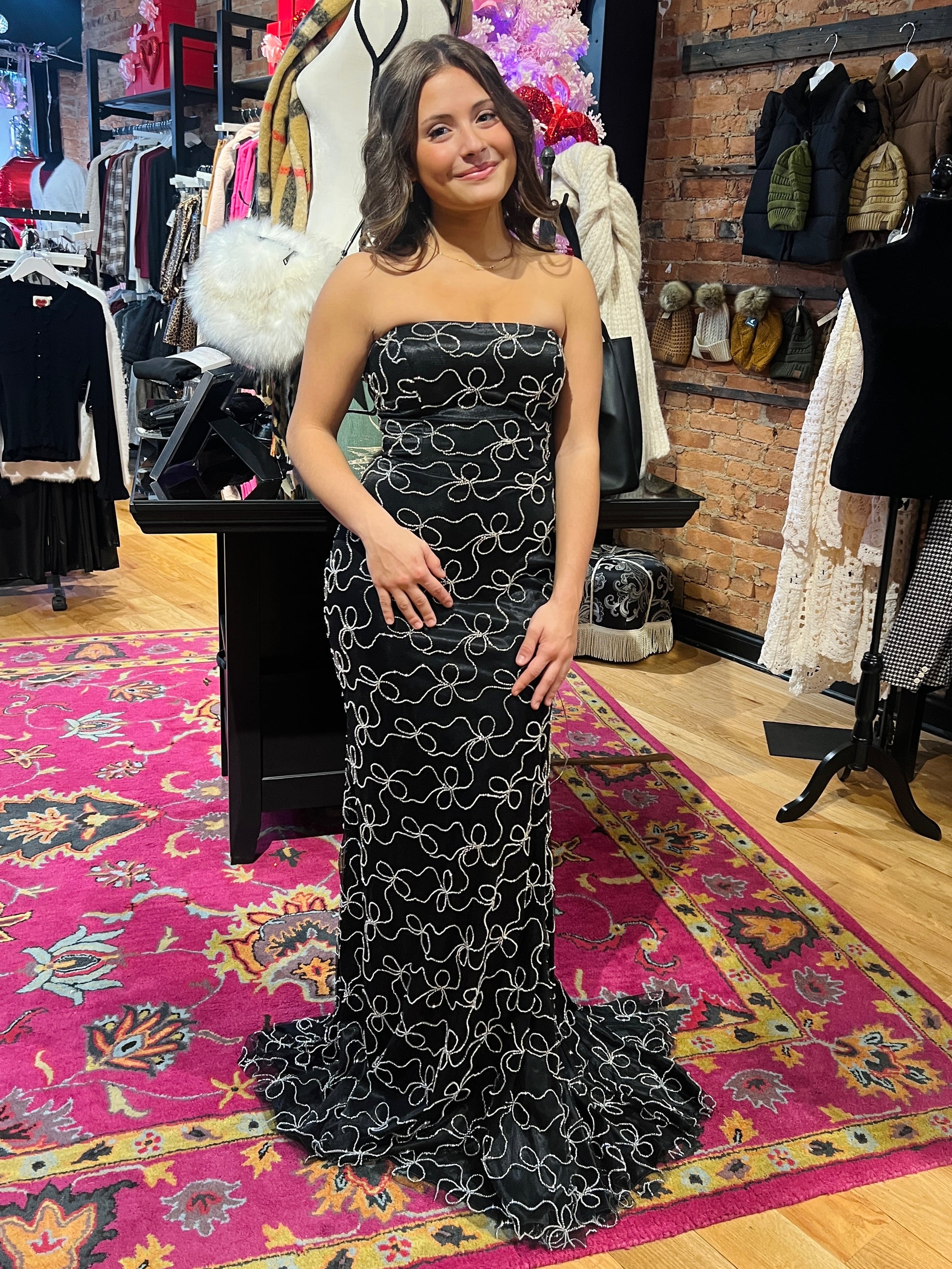 Woman in a strapless black evening gown standing in a clothing store with racks of clothes in the background.