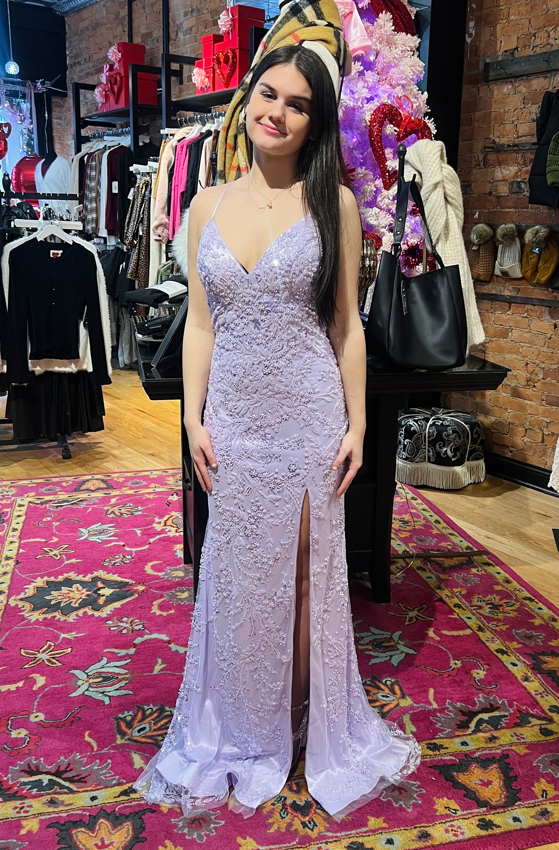 Woman in a silver evening gown standing in a store with racks of clothes and a colorful rug on the floor.