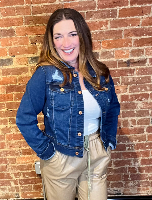 A woman wearing a blue distressed denim jacket with a collared neckline and front button placket, paired with a white top and beige pants, standing against a brick wall.