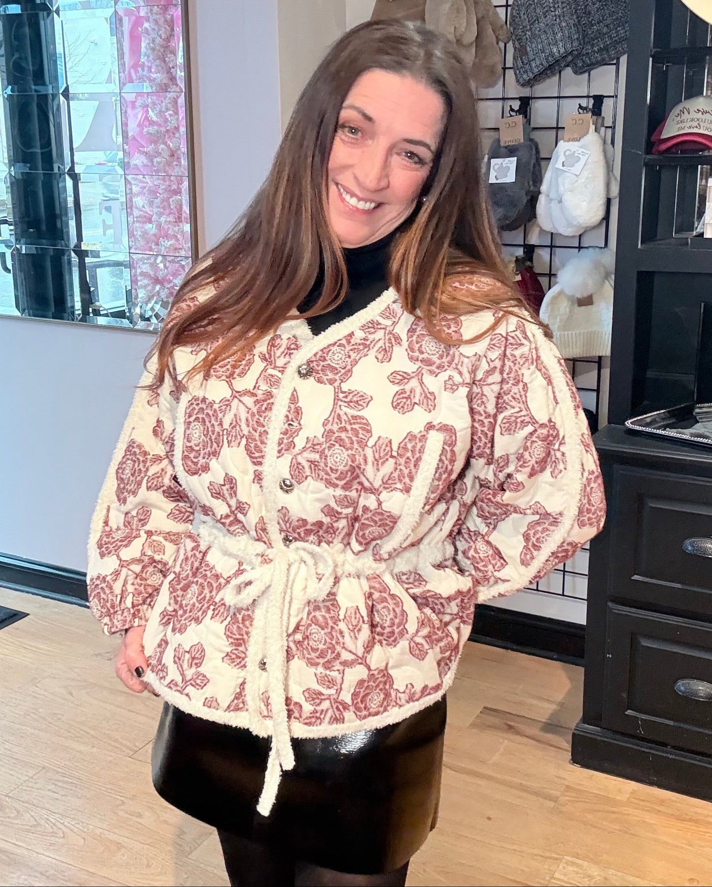 Woman wearing a patterned dress in a store setting with clothing racks and hats.