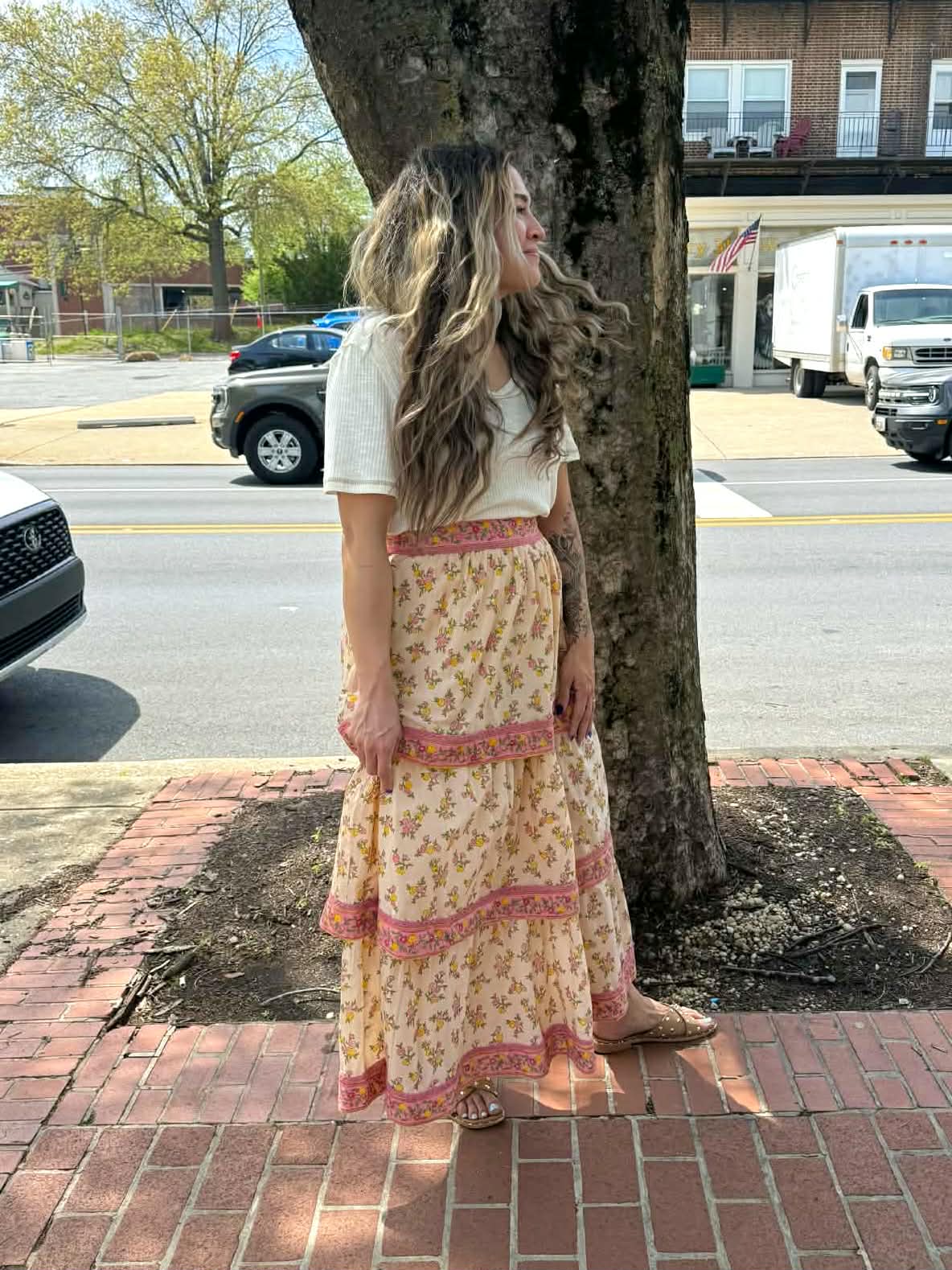 Woman in a long floral dress standing on a sidewalk next to a tree