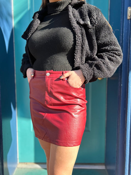 A person wearing a red leather skirt with a black top and a black jacket, standing against a blue background.
