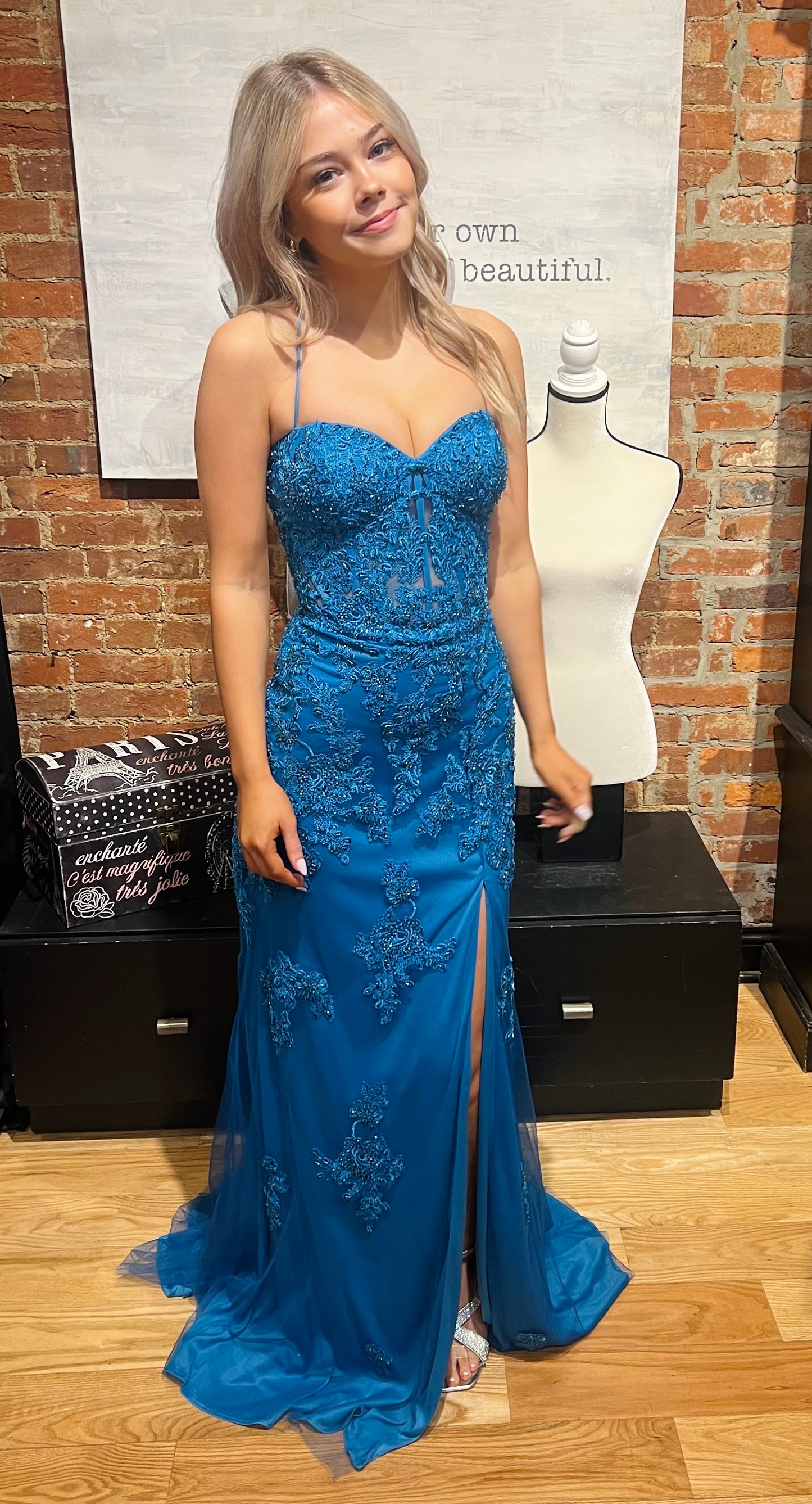 Woman in a blue evening gown standing in front of a brick wall with a mannequin.