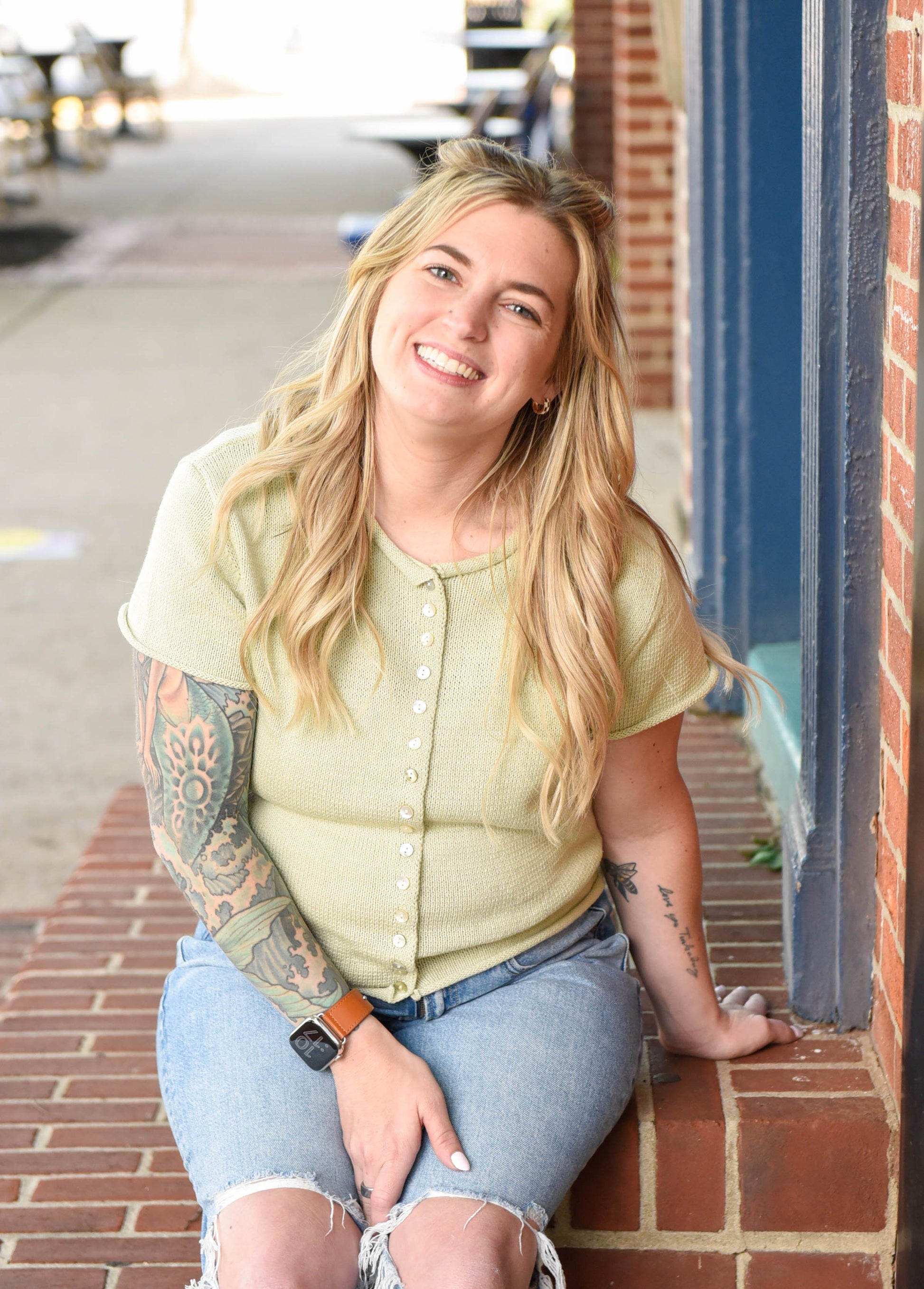 Woman with tattoos wearing a light green shirt and blue jeans sitting on a brick sidewalk.