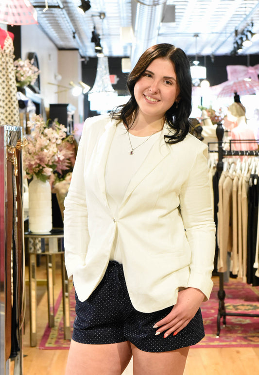 Woman in a white blazer and black shorts standing in a store with racks of clothes and floral arrangements.