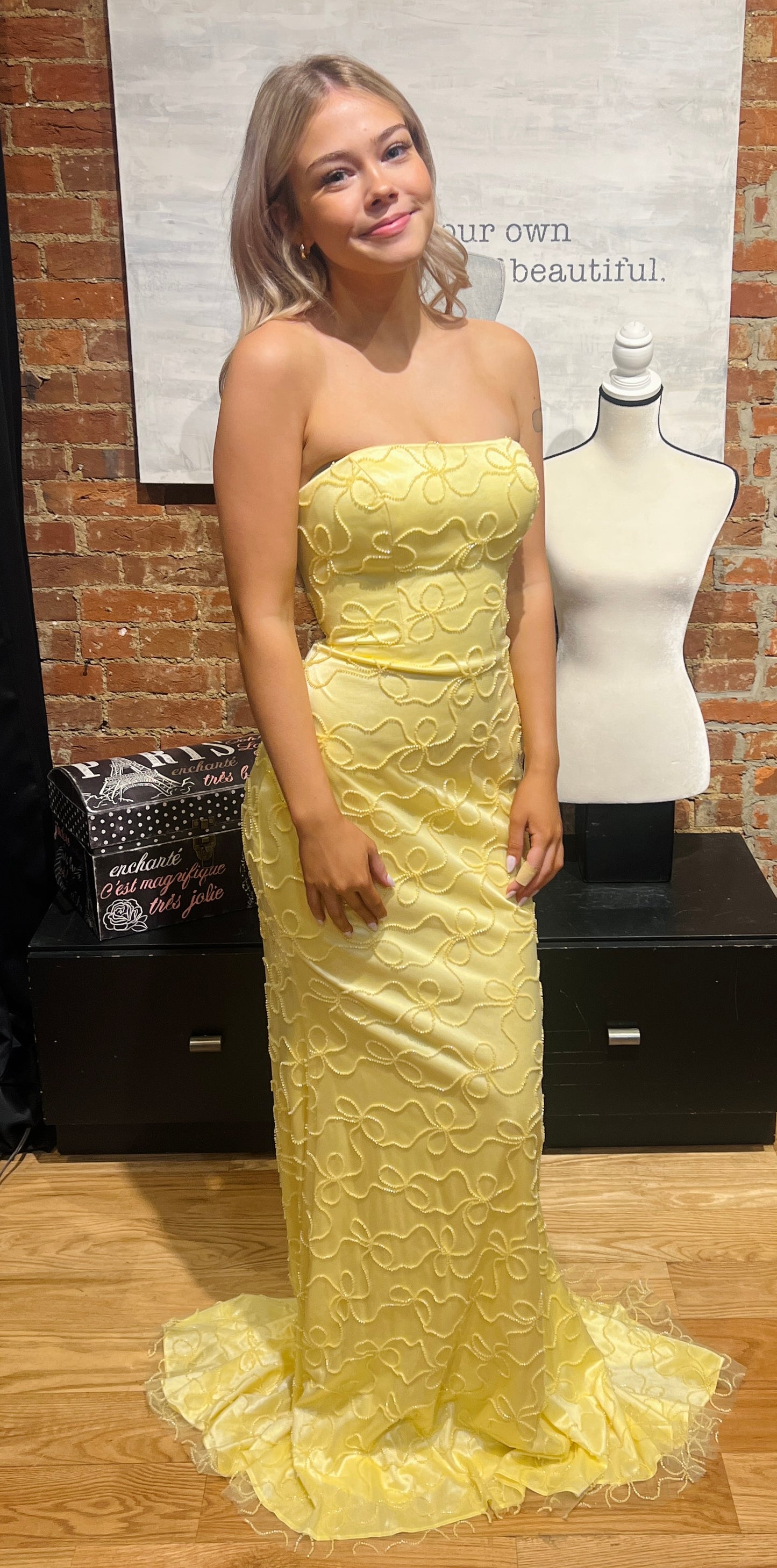Woman in a yellow strapless dress standing next to a mannequin in a room with brick walls.