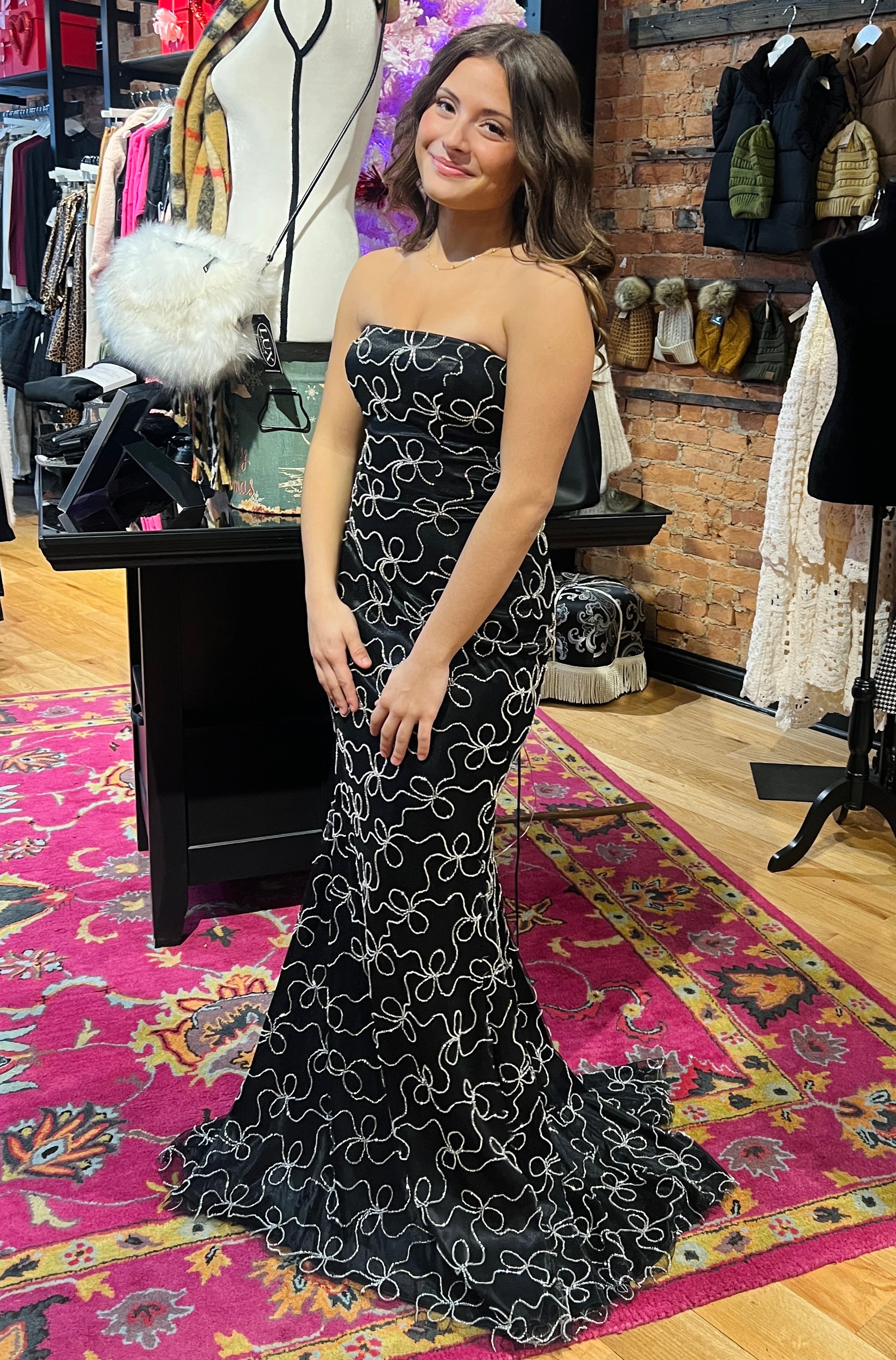 Woman in a black strapless dress with white patterns standing in a store with racks of clothes in the background.