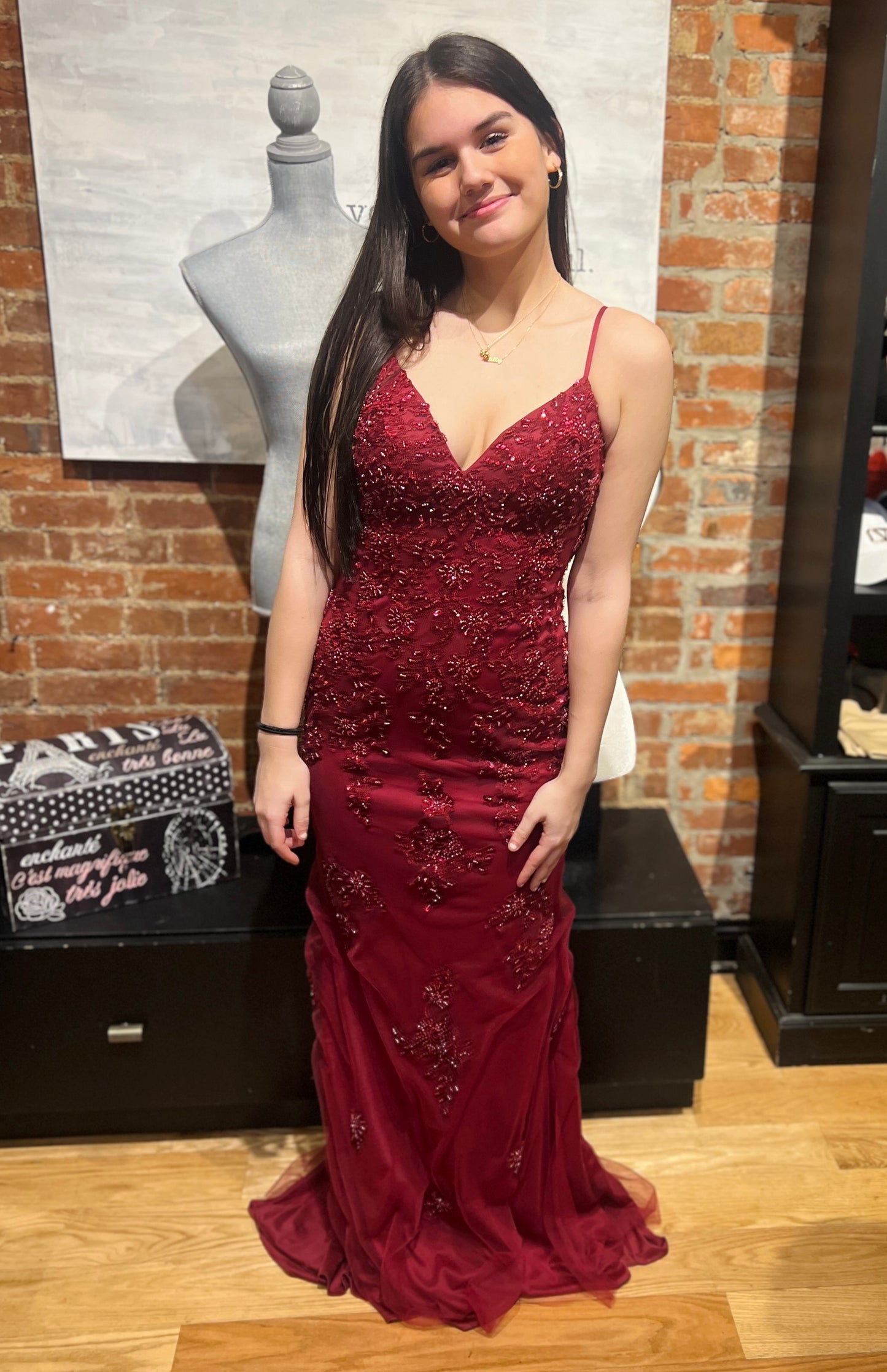 Woman in a burgundy evening gown standing in front of a brick wall with a mannequin and boxes.