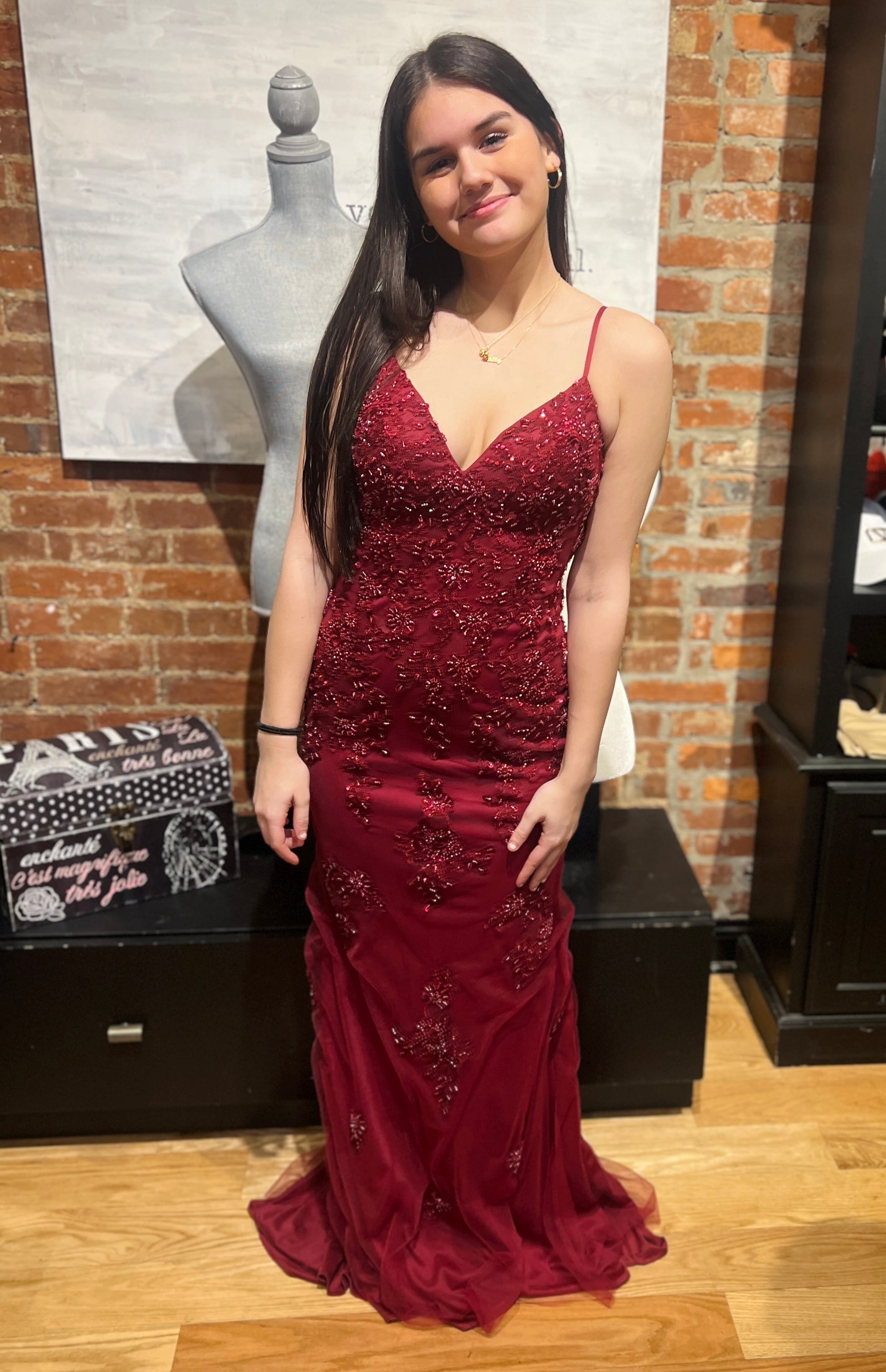 Woman in a burgundy evening gown standing in front of a brick wall with a mannequin and boxes.