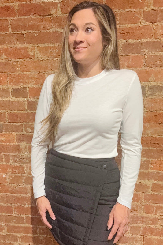 Woman wearing a white long-sleeve top and gray skirt against a brick wall.