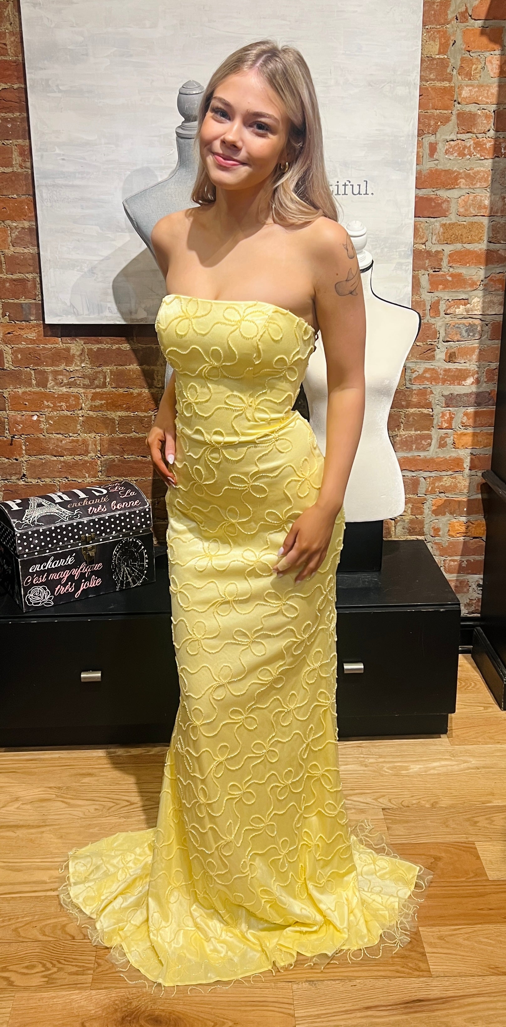 Woman wearing a yellow strapless dress in front of a brick wall.
