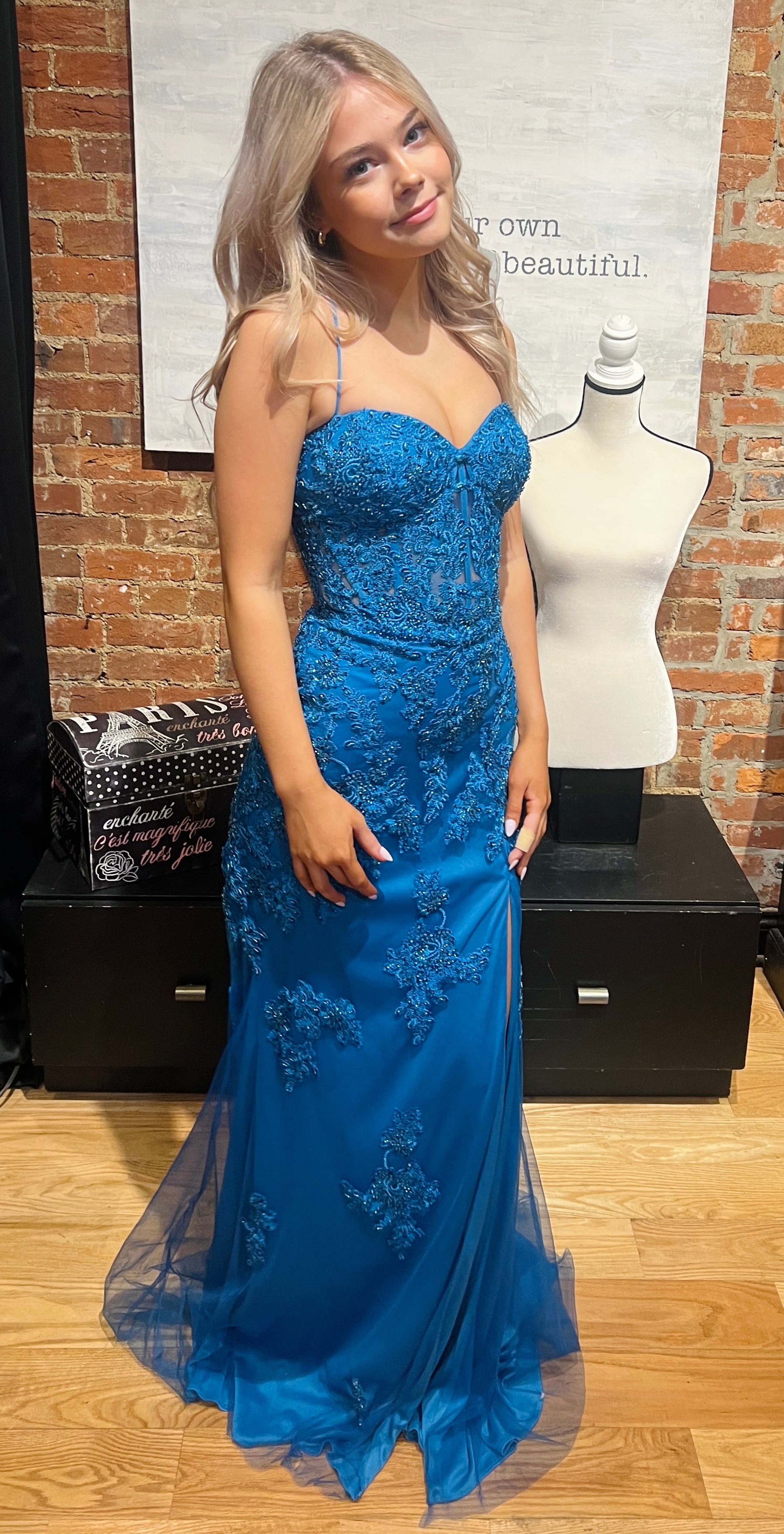 Woman in a blue evening gown standing next to a mannequin with a similar dress against a brick wall.