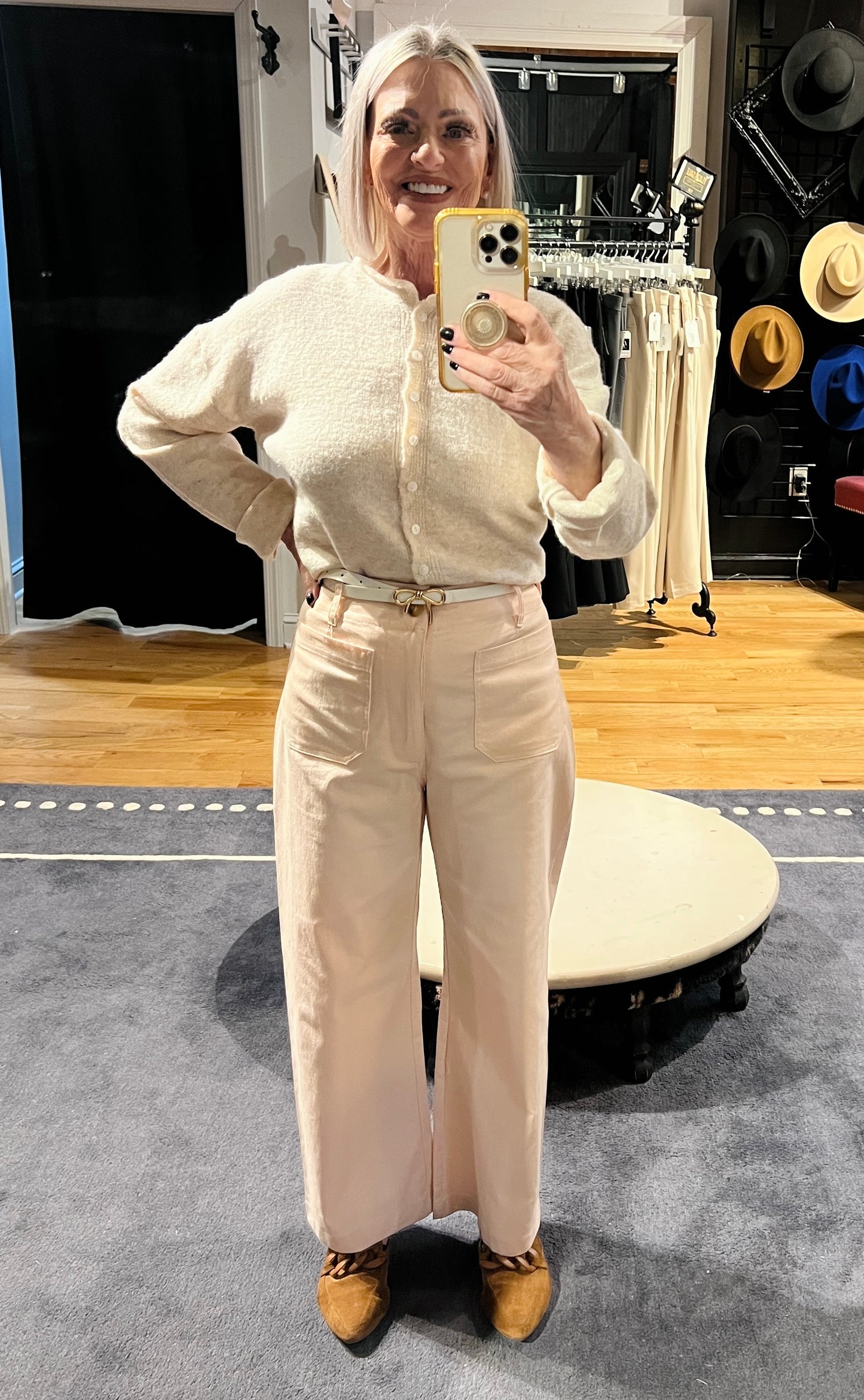 Person wearing a cream outfit taking a mirror selfie in a room with wooden floor and furniture.