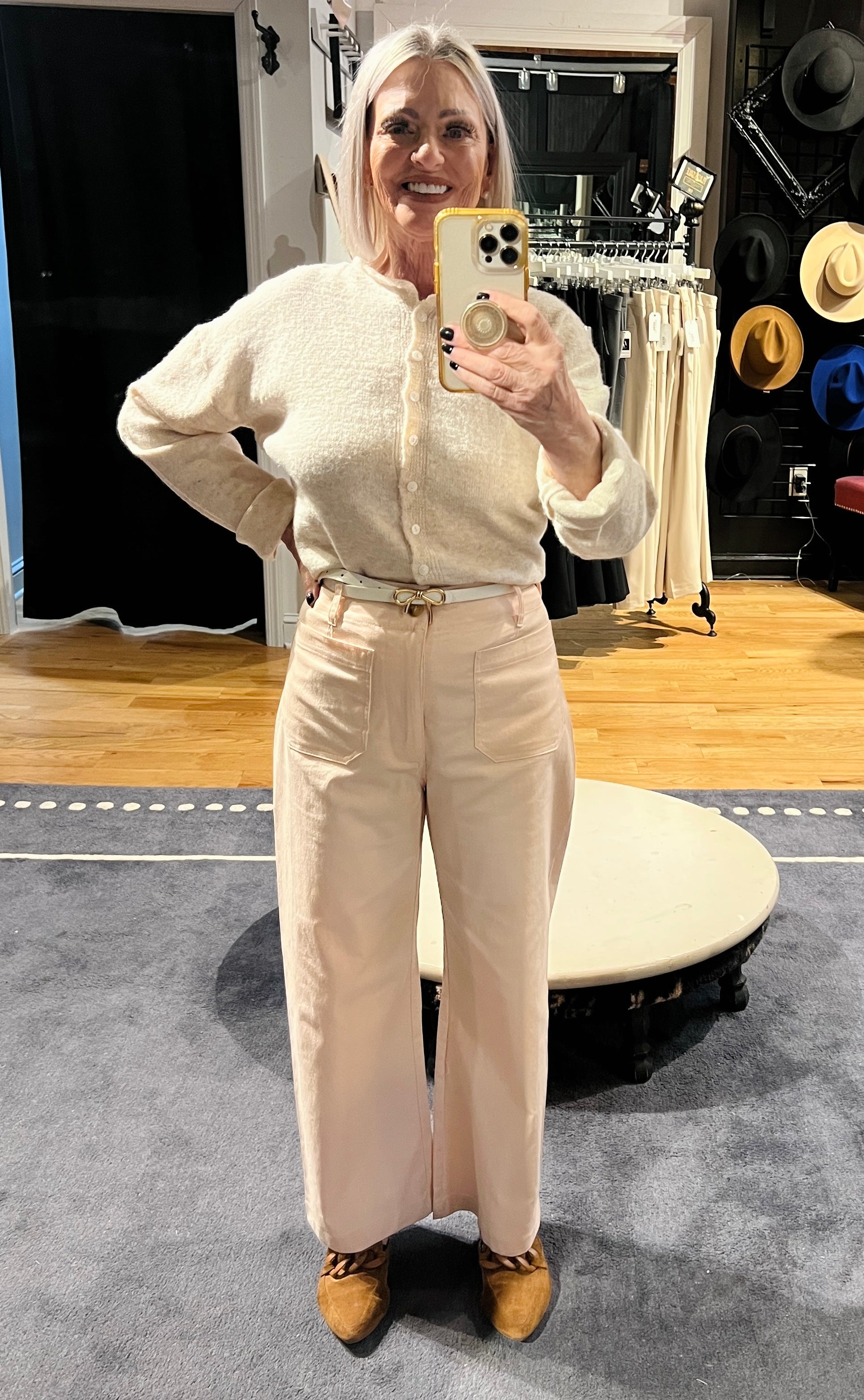 Person wearing a cream outfit taking a mirror selfie in a room with wooden floor and furniture.