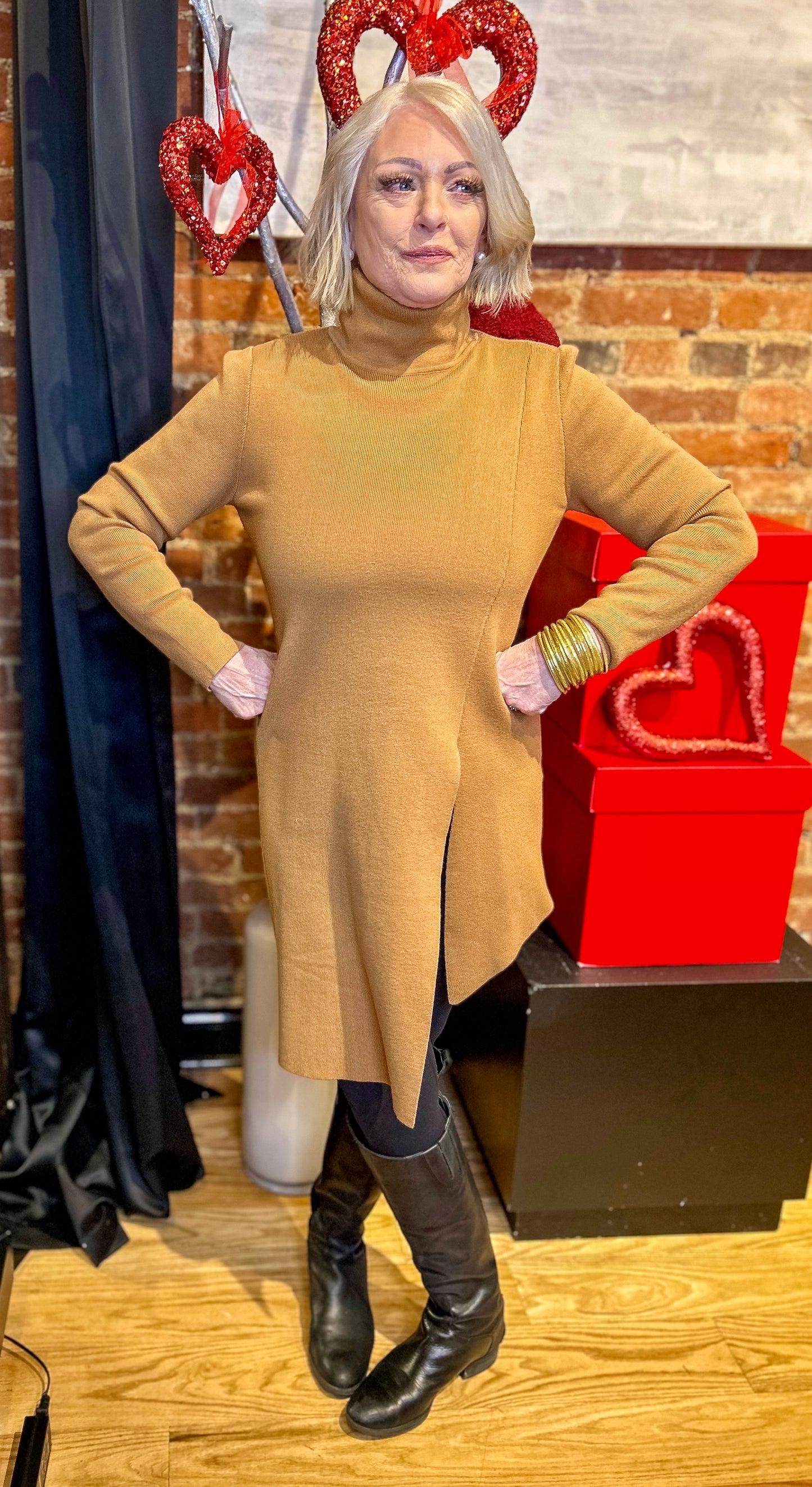 A woman standing indoors wearing a brown asymmetrical hem dress with a turtleneck, paired with black boots and accessorized with red heart-shaped earrings.