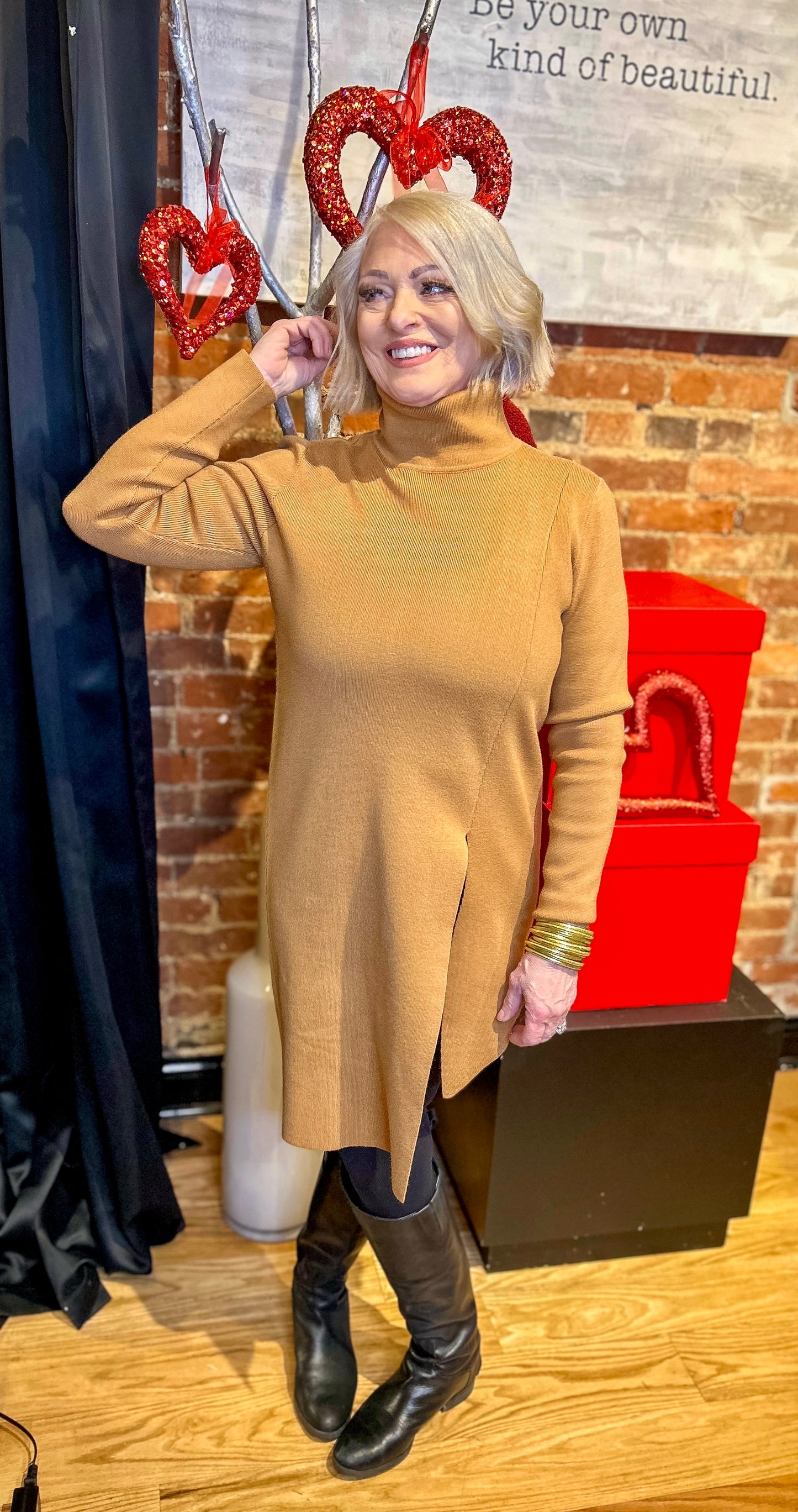 A woman standing indoors wearing a brown asymmetrical hem dress with a turtleneck, paired with black boots and accessorized with red heart-shaped earrings.