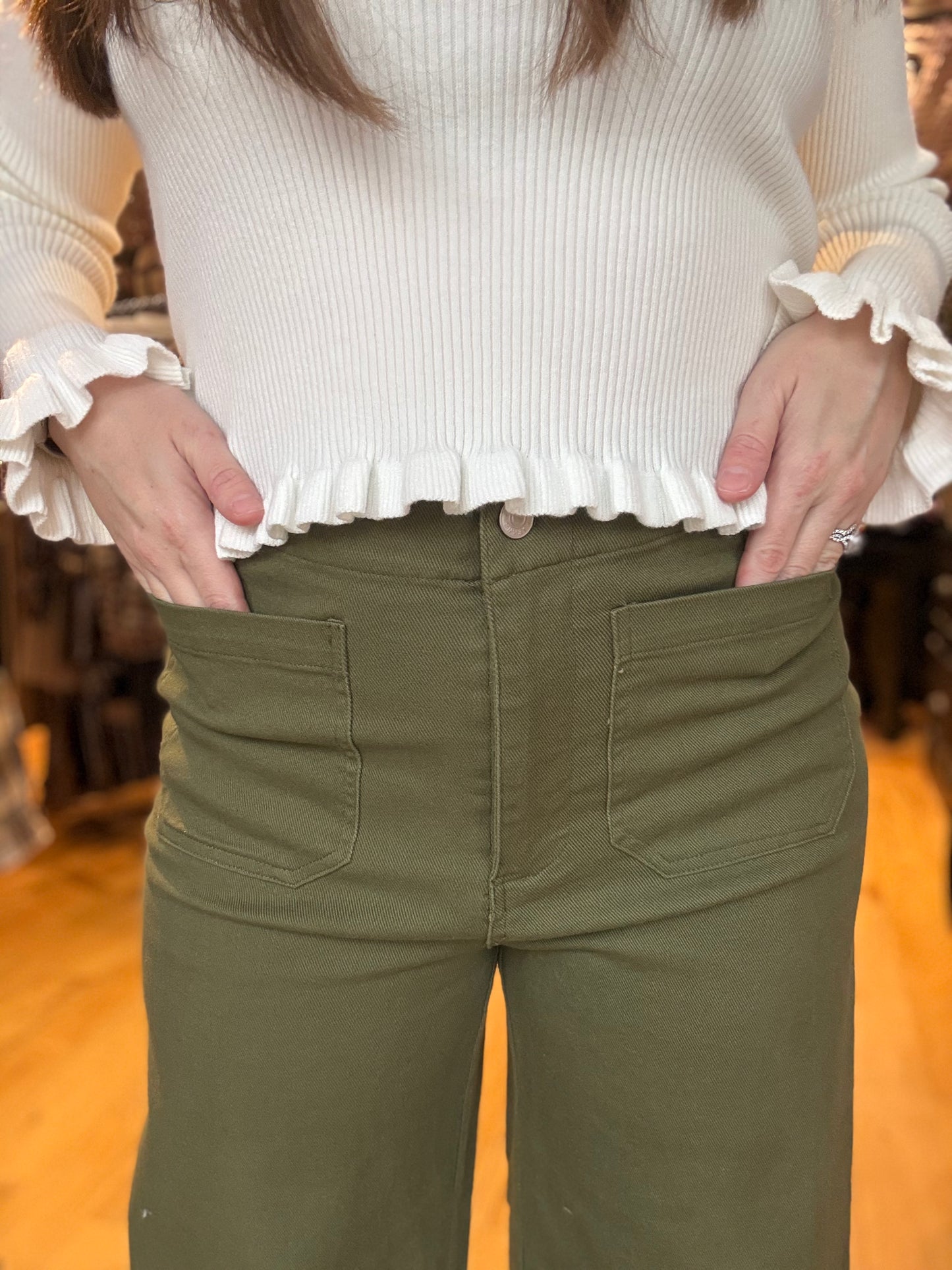 Person wearing a white ruffled blouse and green pants indoors.