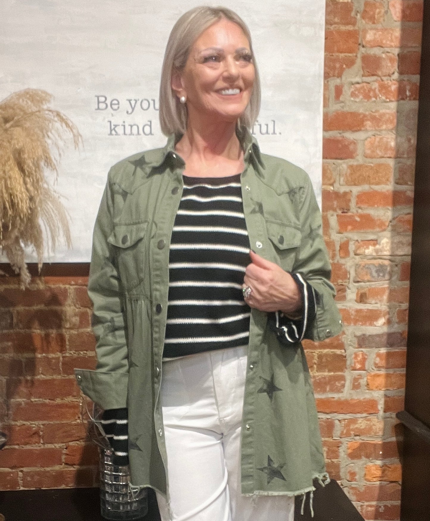 Woman wearing a green jacket with star patterns, black and white striped shirt, and white pants standing against a brick wall.