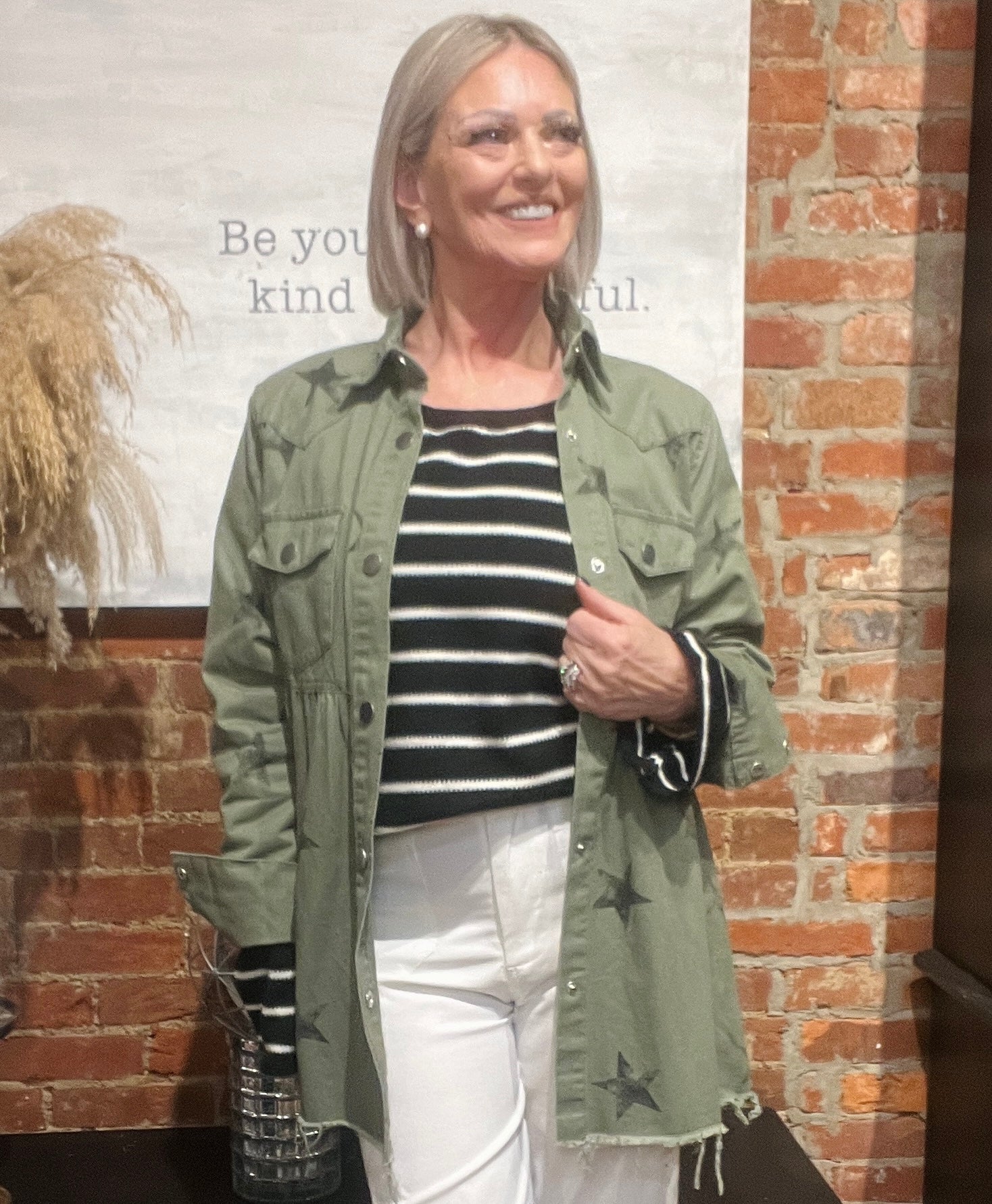 Woman wearing a green jacket with star patterns, black and white striped shirt, and white pants standing against a brick wall.
