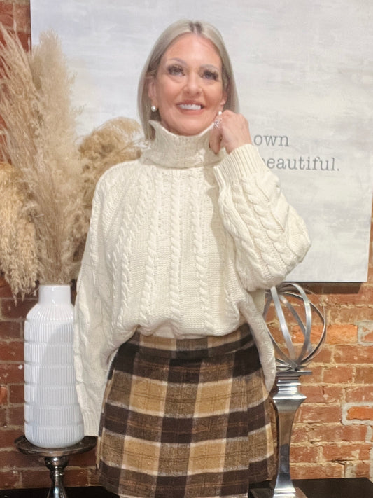 Woman wearing a cream sweater and plaid skirt in a decorative setting with a vase and decor items.