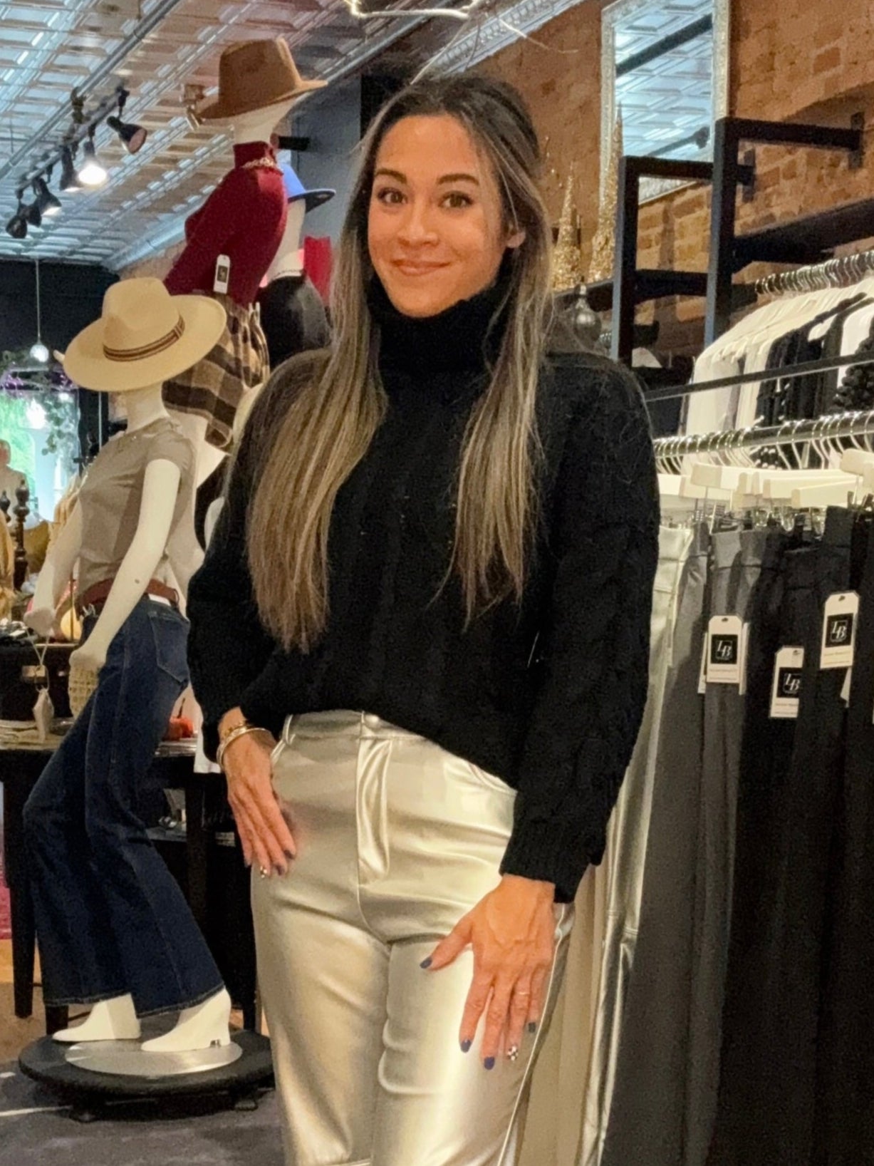 Woman in a store wearing a black top and white pants, surrounded by clothing racks.