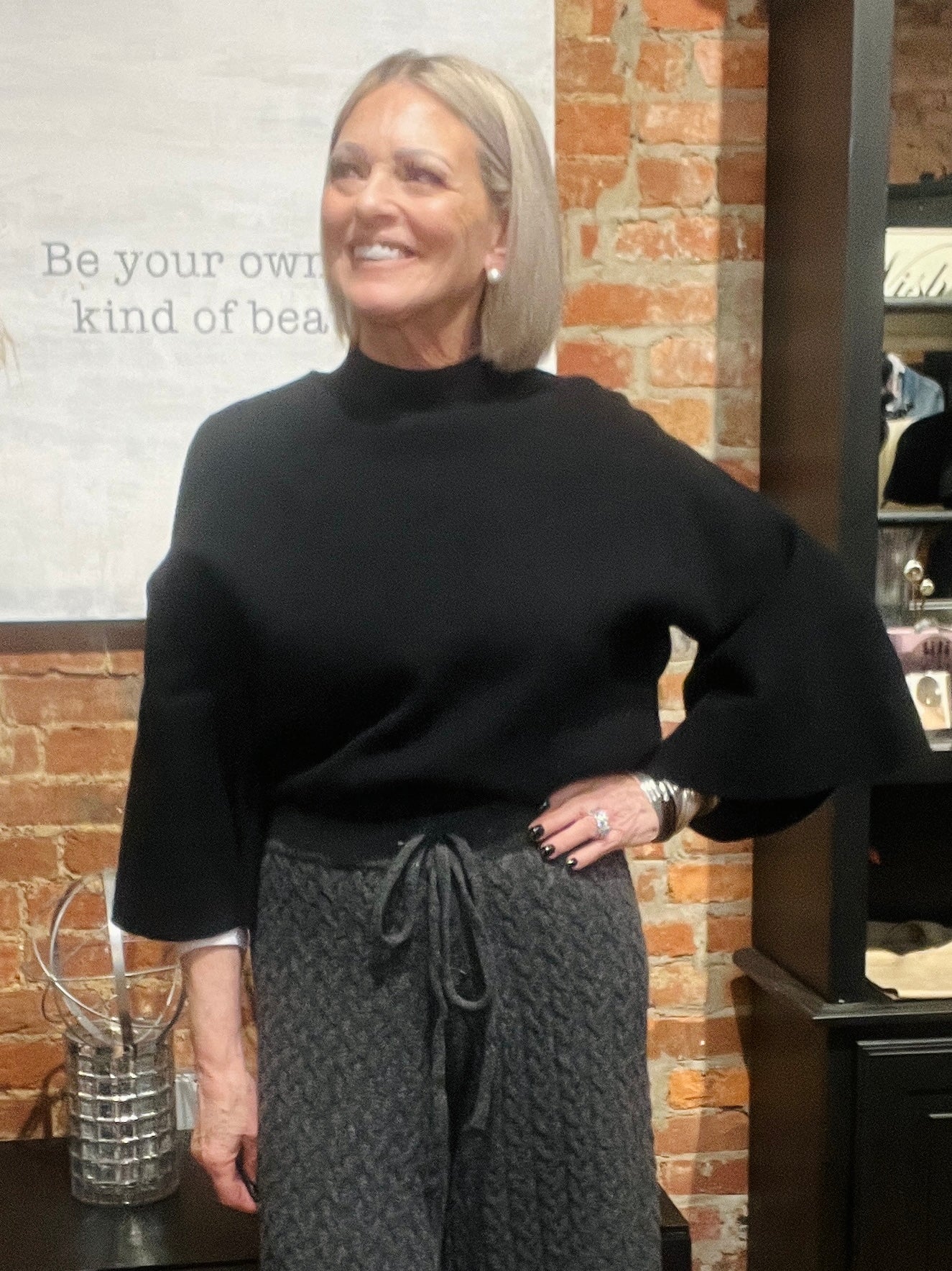 Woman wearing a black sweater and gray pants standing in front of a brick wall.