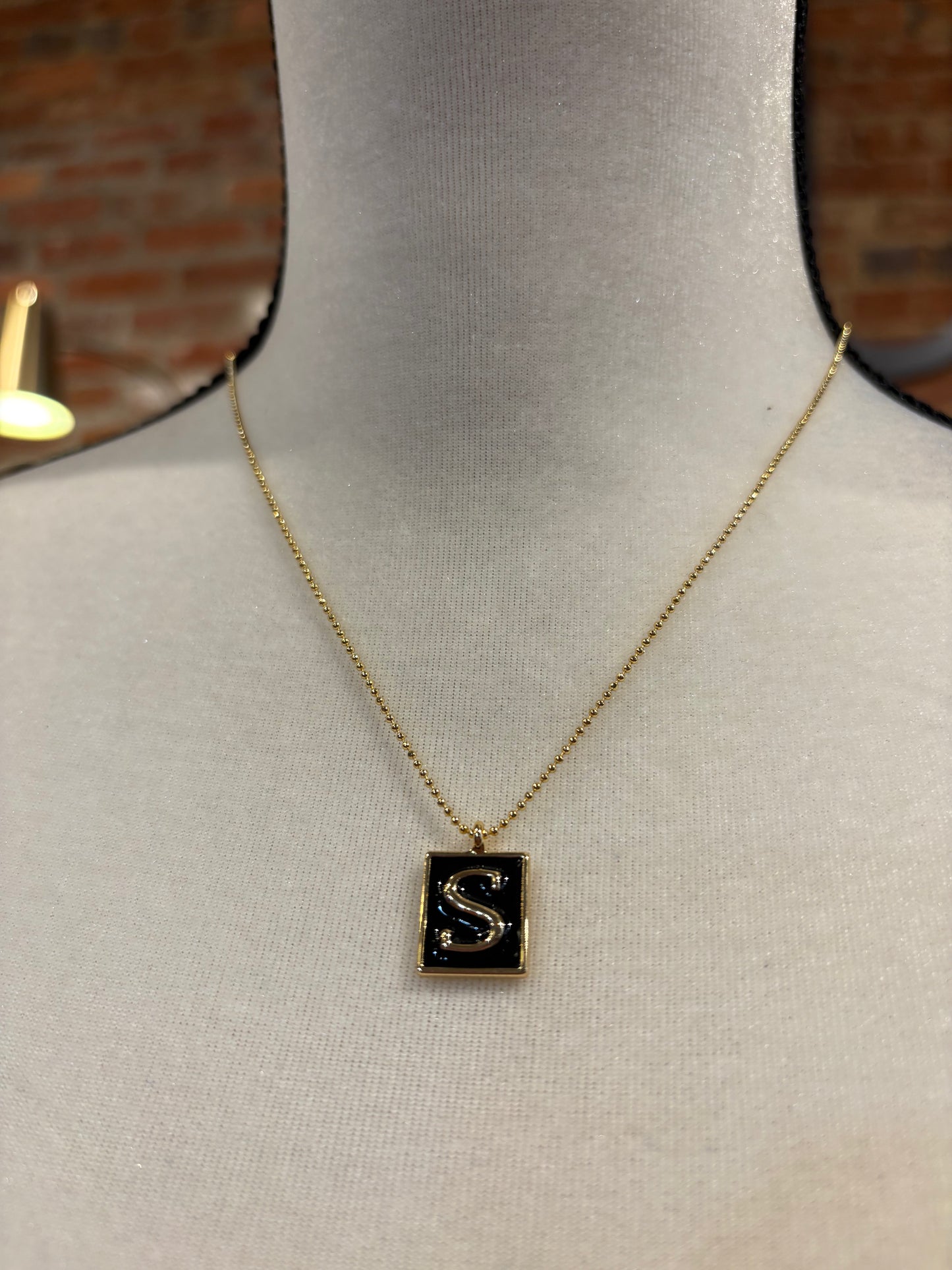 Necklace with a pendant featuring the letter 'S' on a mannequin against a brick wall.