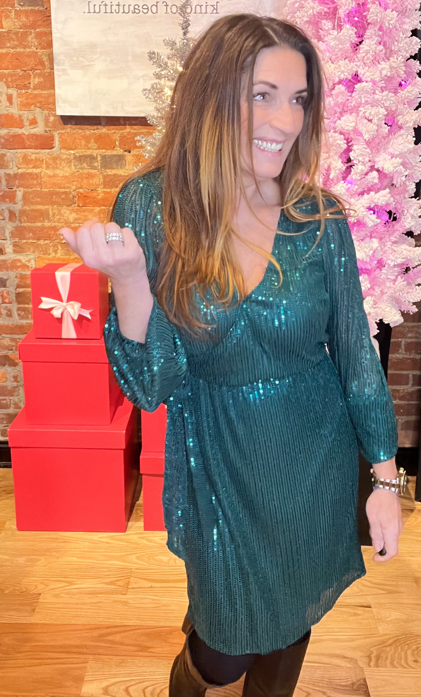 A woman standing in a holiday wrap dress with sequin design, v-neckline, and tie waist, smiling and posing by a Christmas tree and gifts.