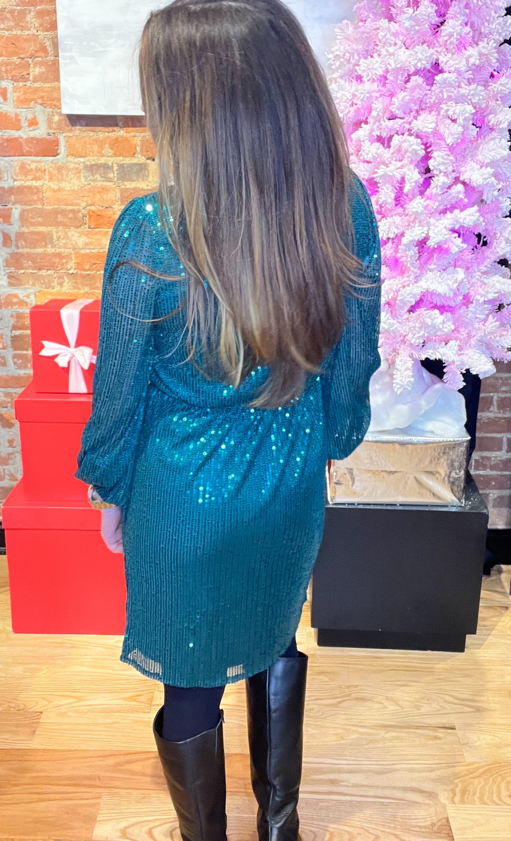 A woman standing in a holiday wrap dress with sequin design, v-neckline, and tie waist, smiling and posing by a Christmas tree and gifts.