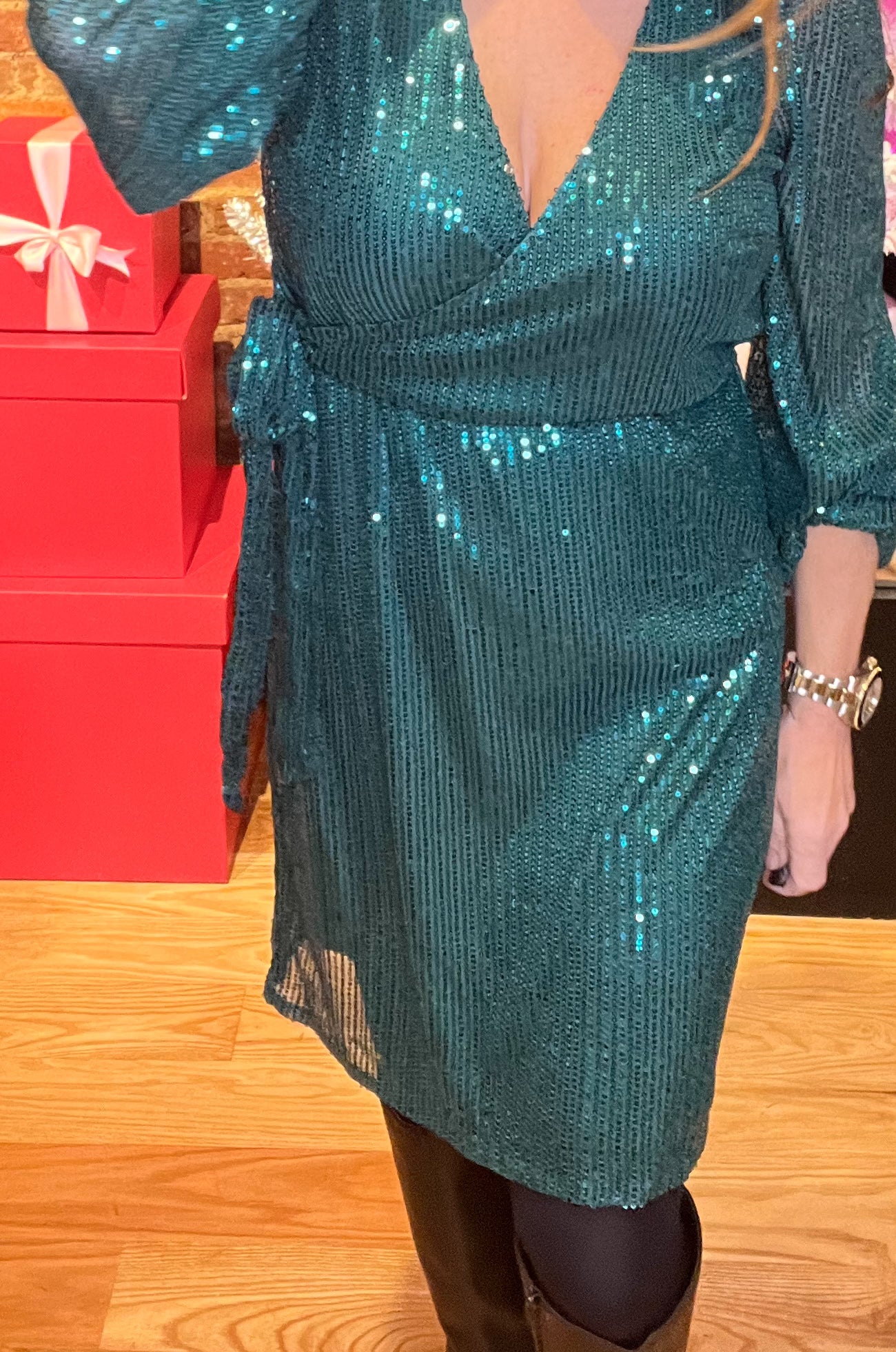 A woman standing in a holiday wrap dress with sequin design, v-neckline, and tie waist, smiling and posing by a Christmas tree and gifts.