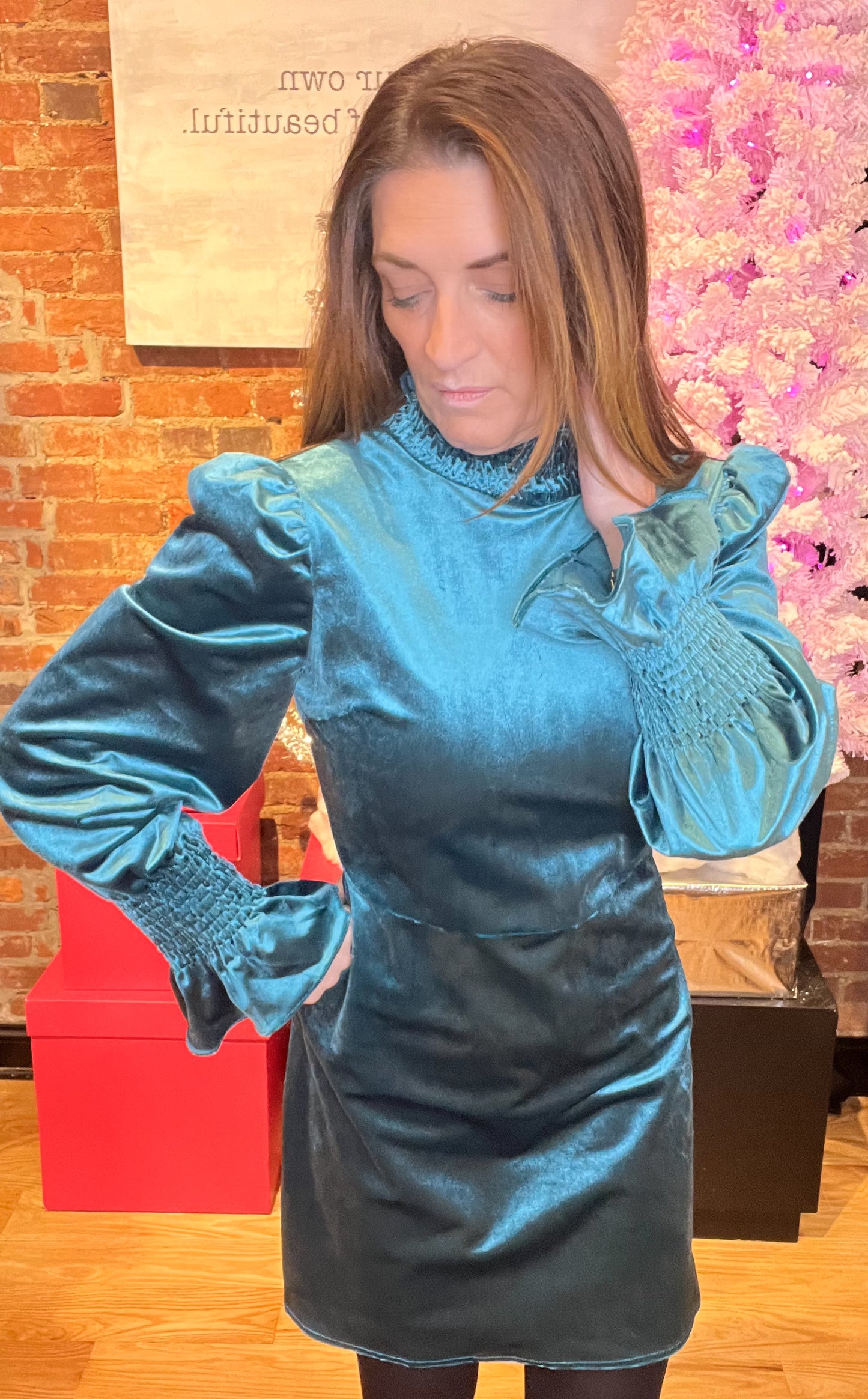 A woman wearing a teal velvet mini dress with a smocked neck and sleeves, paired with black tights and a pair of shoes.