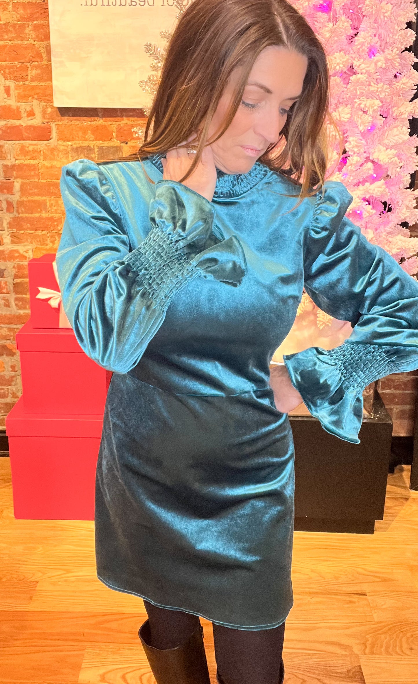 A woman wearing a teal velvet mini dress with a smocked neck and sleeves, paired with black tights and a pair of shoes.