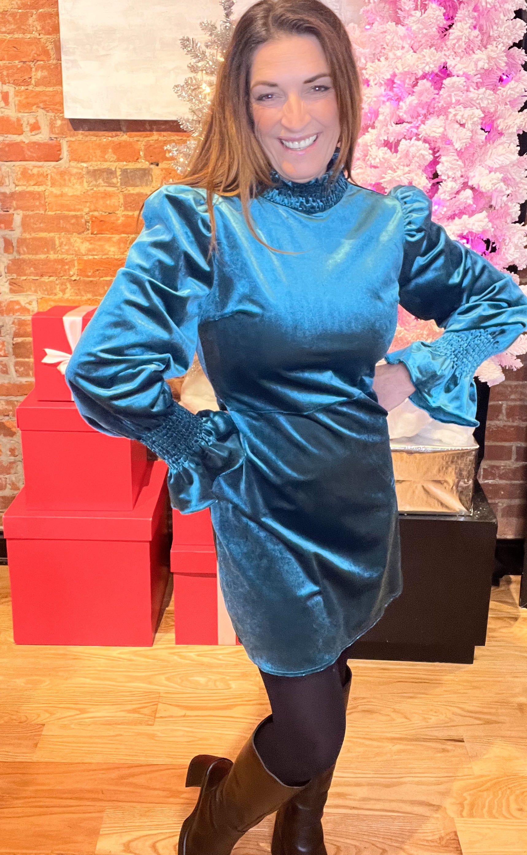 A woman wearing a teal velvet mini dress with a smocked neck and sleeves, paired with black tights and a pair of shoes.