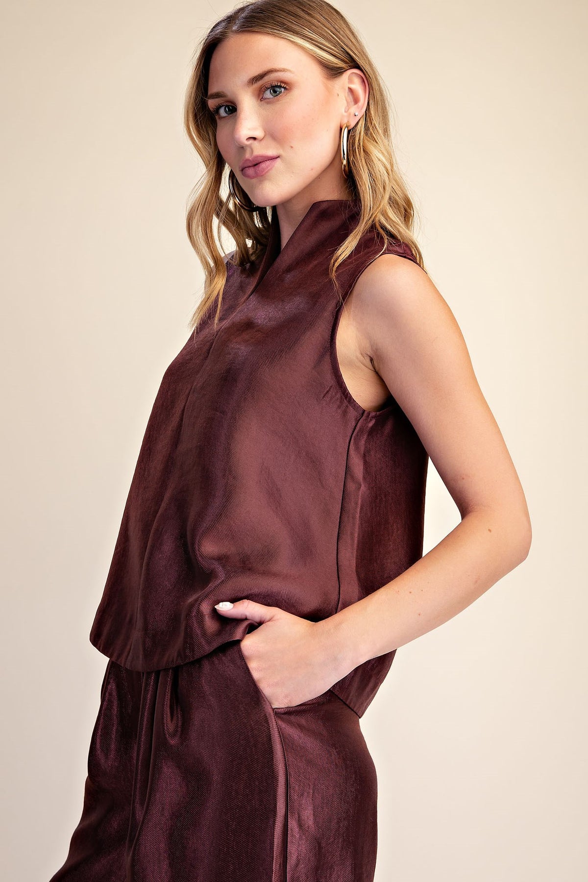 A woman modeling a sleeveless high-neck top in a deep burgundy or maroon shade, paired with matching bottoms, against a plain beige studio background.