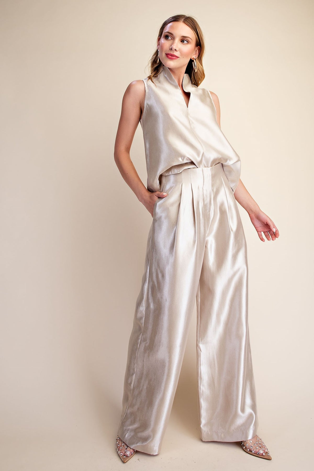 A woman modeling a sleeveless high-neck jumpsuit in a shiny champagne-colored satin fabric, set against a plain beige studio background.