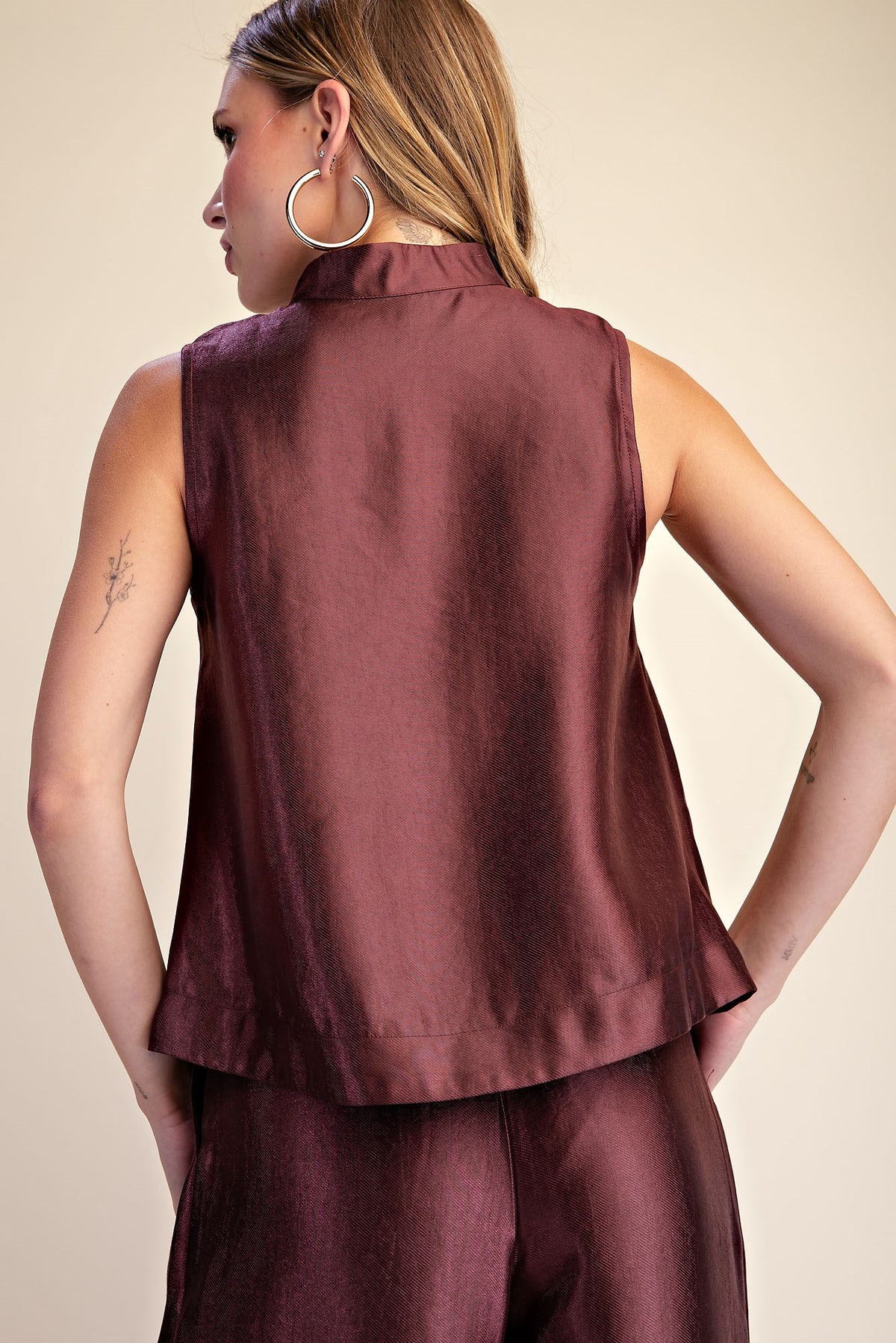 A woman shown from the back wearing a sleeveless, high-neck top and matching pants in a deep burgundy or maroon satin-like fabric, set against a plain beige studio background.