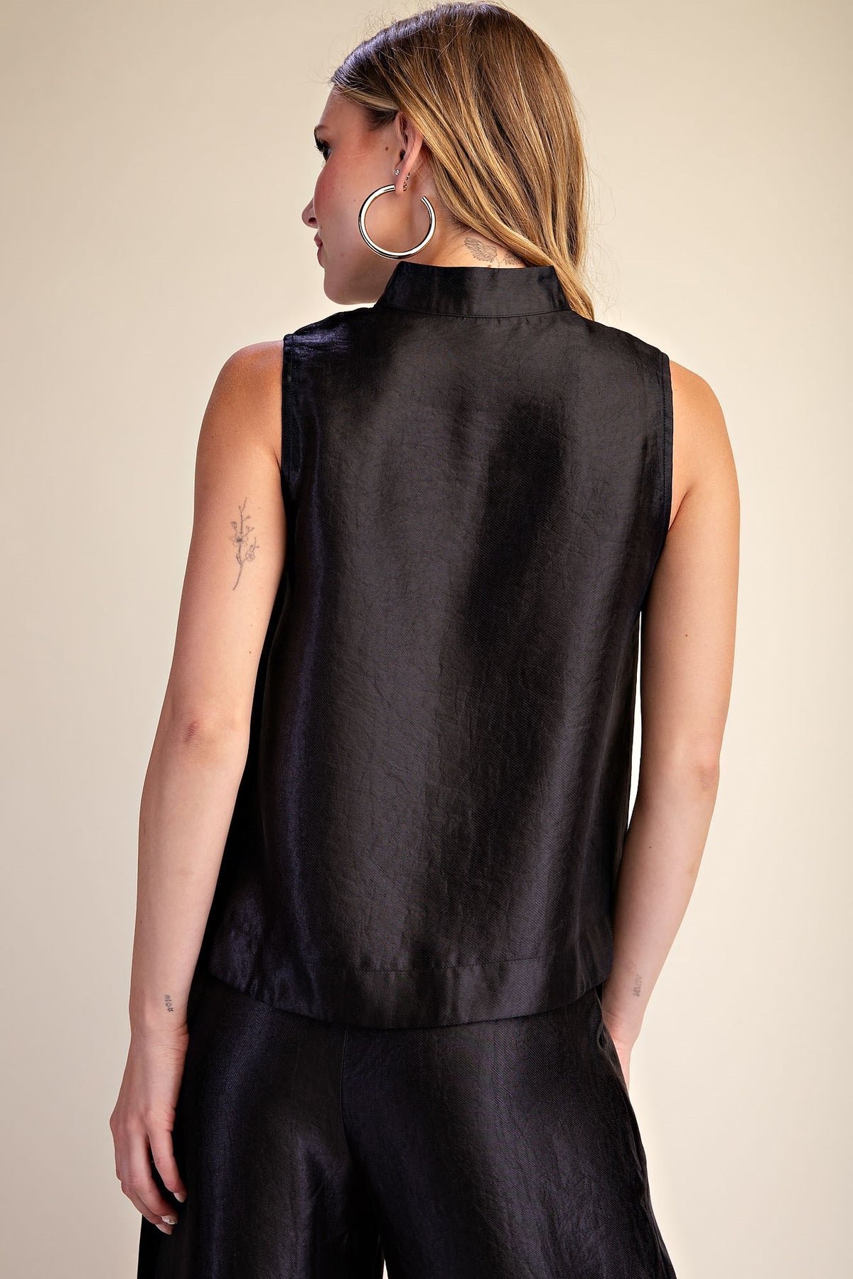 A woman shown from the back wearing a sleeveless black satin-like top and matching black bottoms against a plain beige studio background.