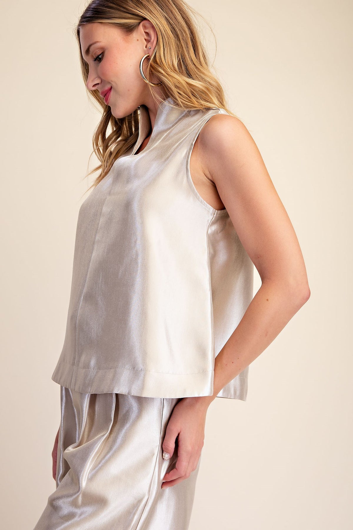 A woman wearing a sleeveless, high-neck satin top and matching pants in a light champagne color, set against a plain beige background.
