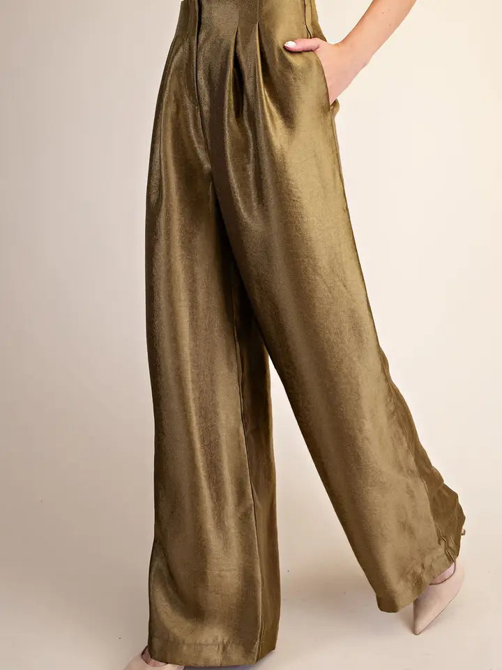 A close-up of a person wearing high-waisted, wide-leg trousers in a shiny olive-gold fabric, with one hand in a pocket, against a plain beige studio background.