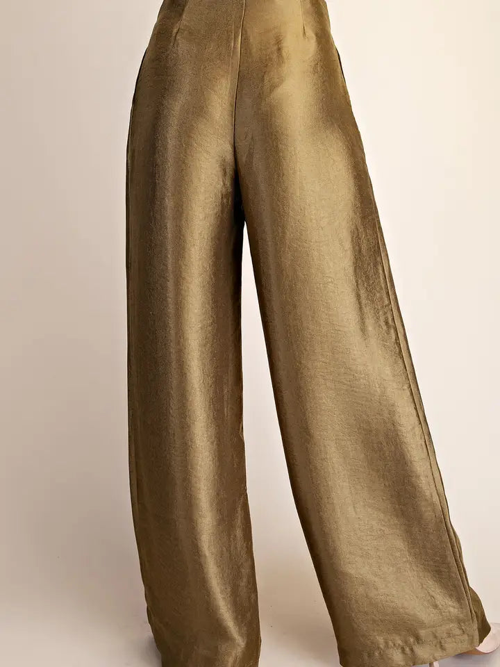 A close-up of a person wearing wide-leg trousers in a shiny olive-gold fabric, shown from the waist down against a plain light beige studio background.