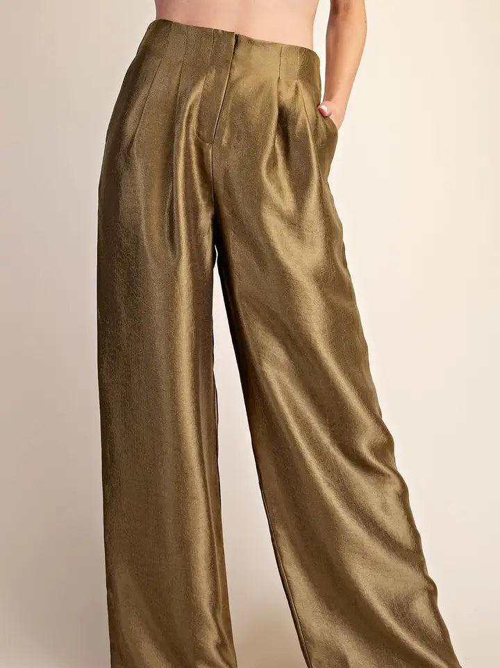 A close-up of a model wearing high-waisted, wide-leg trousers in a shimmering metallic olive-gold fabric, with one hand in a pocket, set against a plain beige background.