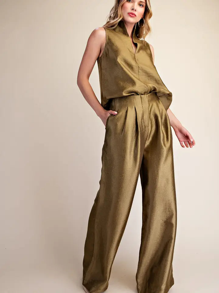 A woman modeling a sleeveless olive green jumpsuit with a high-waisted, wide-leg design and a satin-like sheen, set against a plain beige studio background.