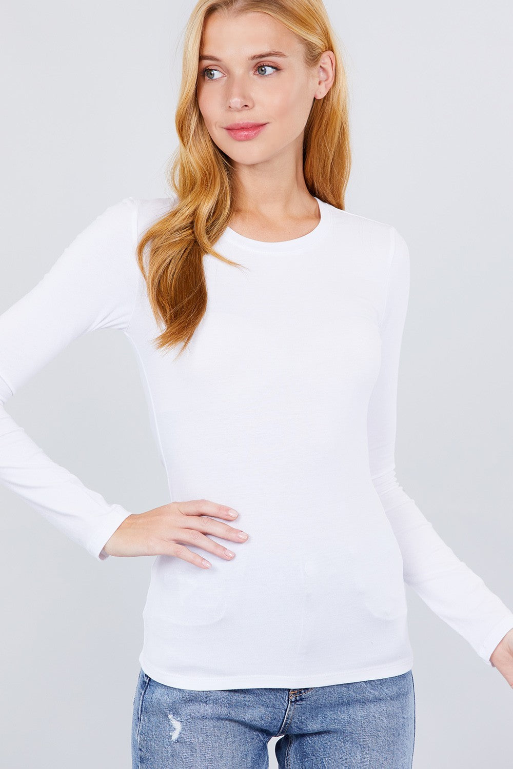 A woman wearing a fitted white long-sleeve crewneck top and blue jeans, standing against a light gray studio background.