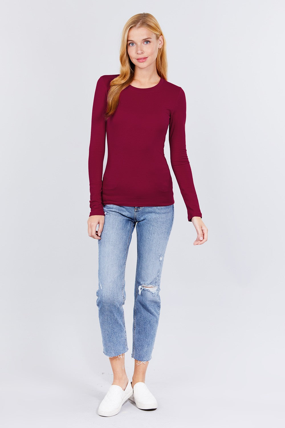 A woman modeling a long-sleeve crewneck sweater in deep burgundy, paired with light-wash distressed jeans and white slip-on shoes, against a plain light gray studio background.