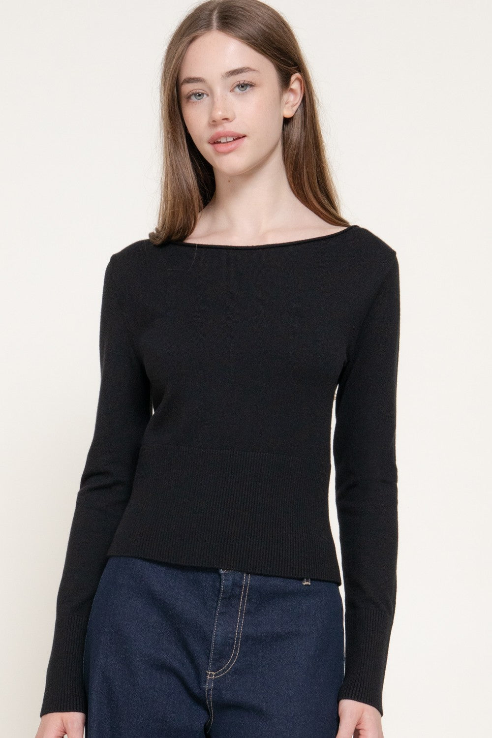 A woman modeling a black long-sleeve knit top with a wide boat neckline, paired with dark blue jeans, against a plain off-white studio background.